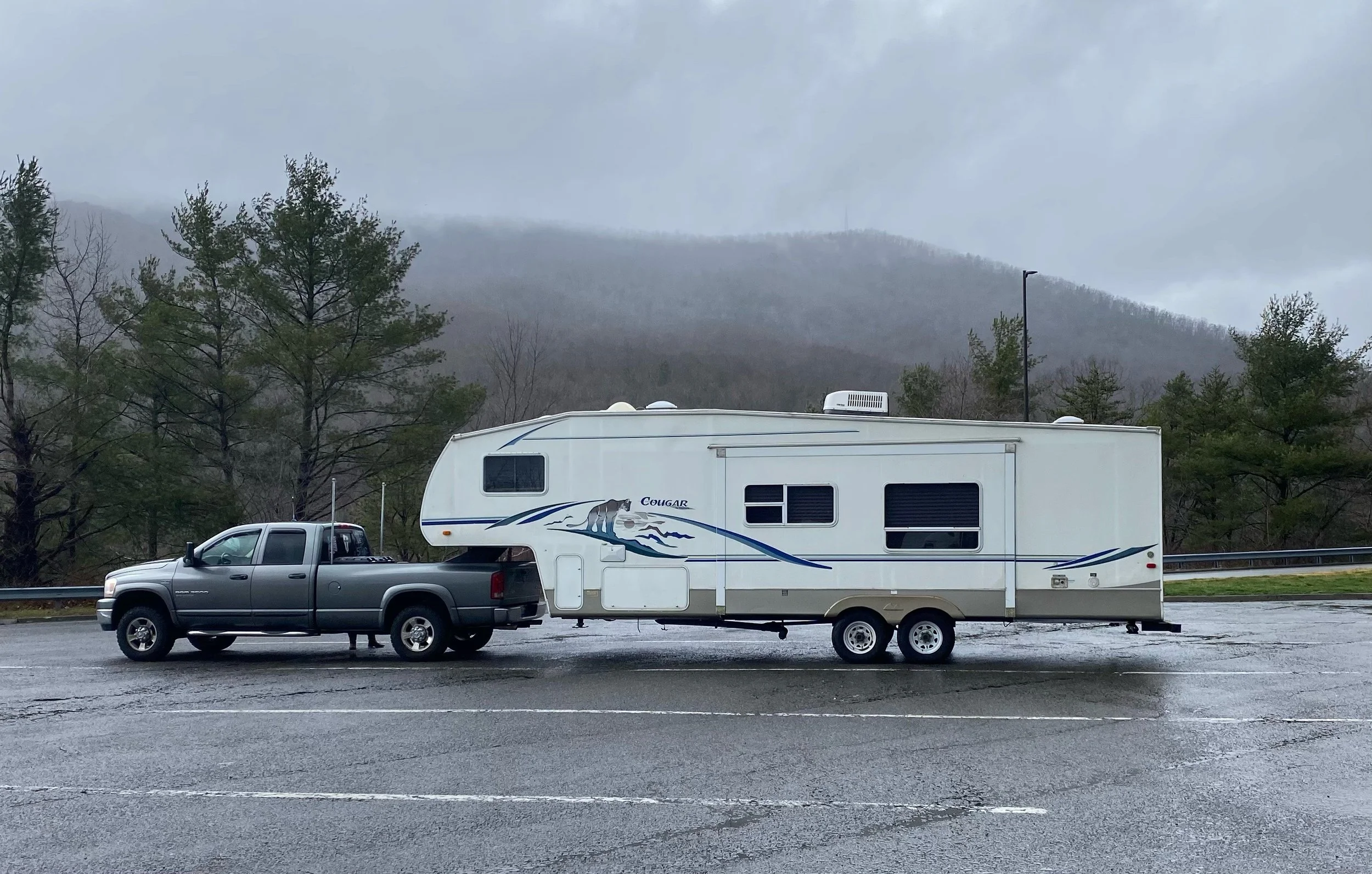 Gray pickup truck towing a white travel trailer with mountain and trees in background on a rainy day.