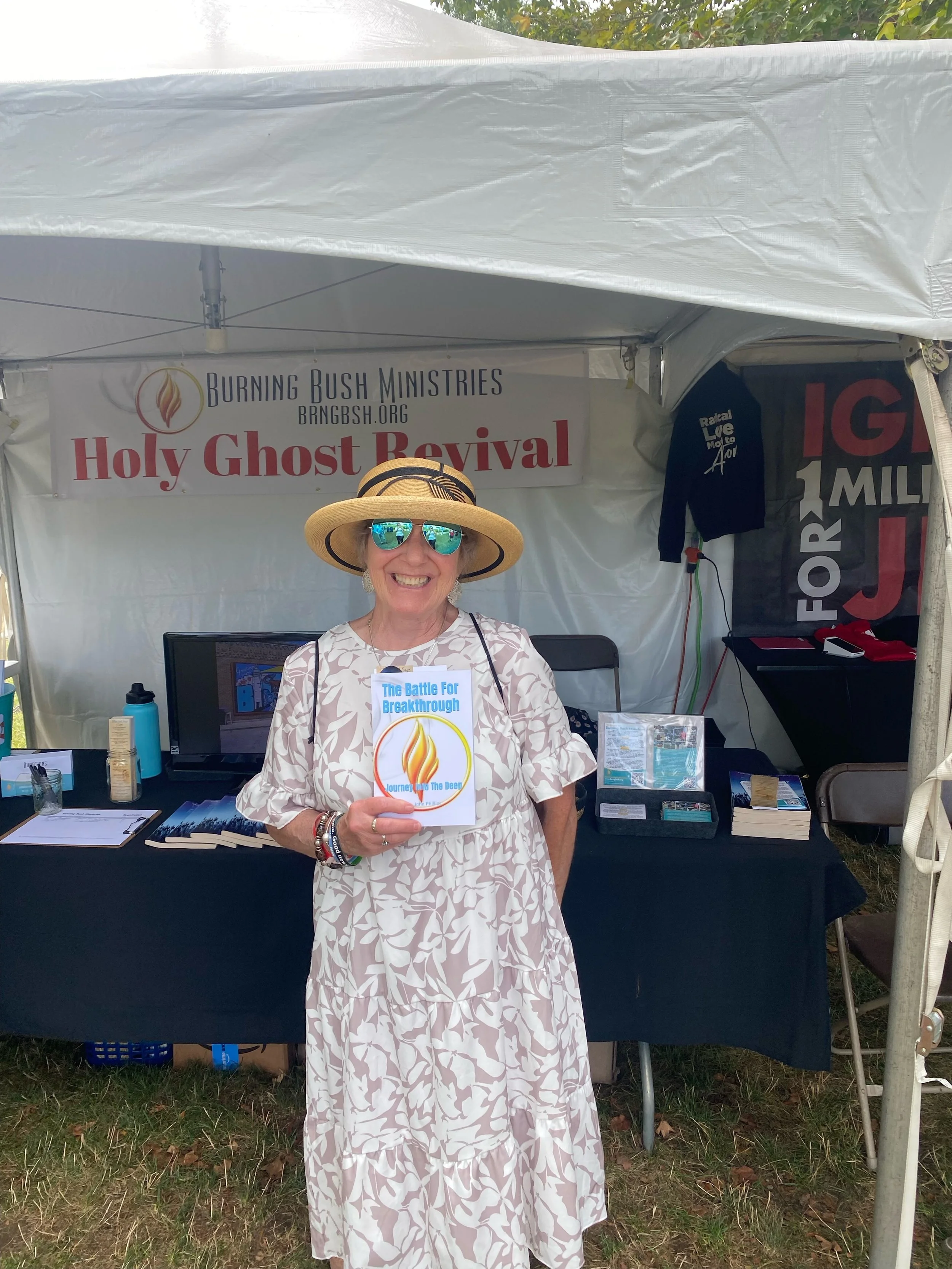 Smiling woman in a white patterned dress and large hat standing in front of a booth with signs for Holy Ghost Revival and Burning Bush Ministries, holding a pamphlet titled 'The Battle For Breakthrough'