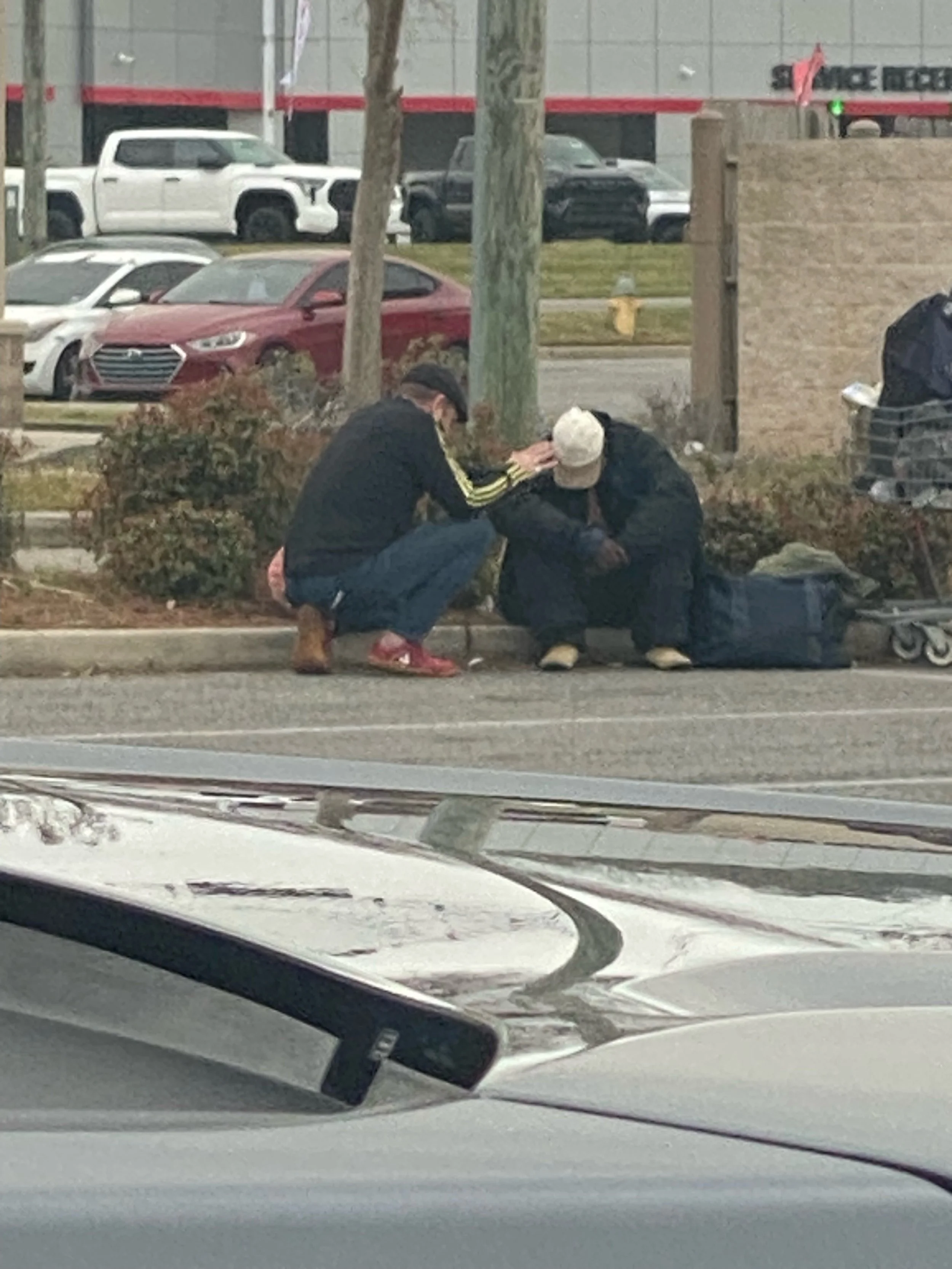 Two men sitting on a sidewalk curb, one praying with the other, outside a parking lot with cars and trees in the background.