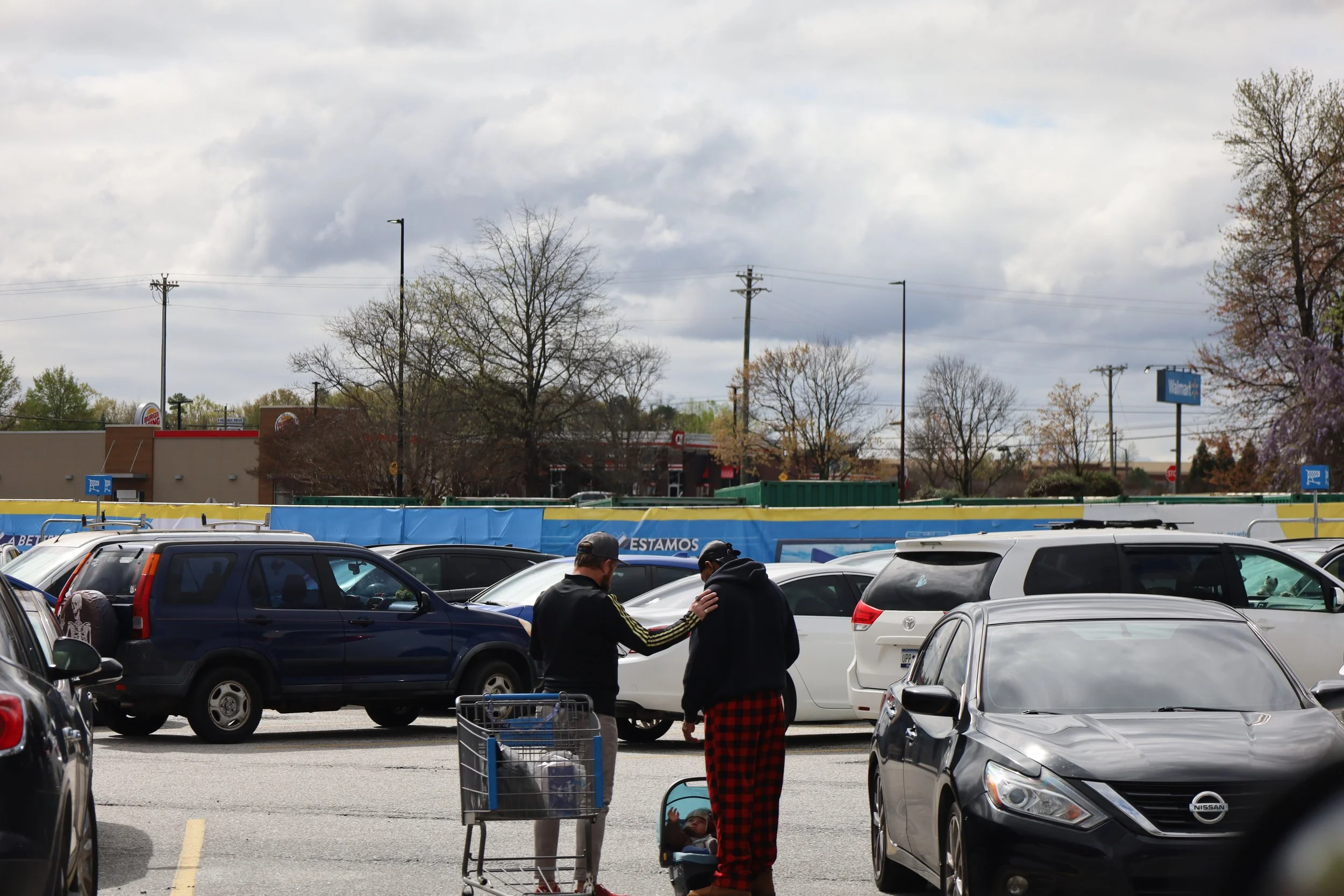 Two people standing in a parking lot, one with a shopping cart, engaged in conversation and prayer amidst parked cars, with trees and commercial buildings in the background.