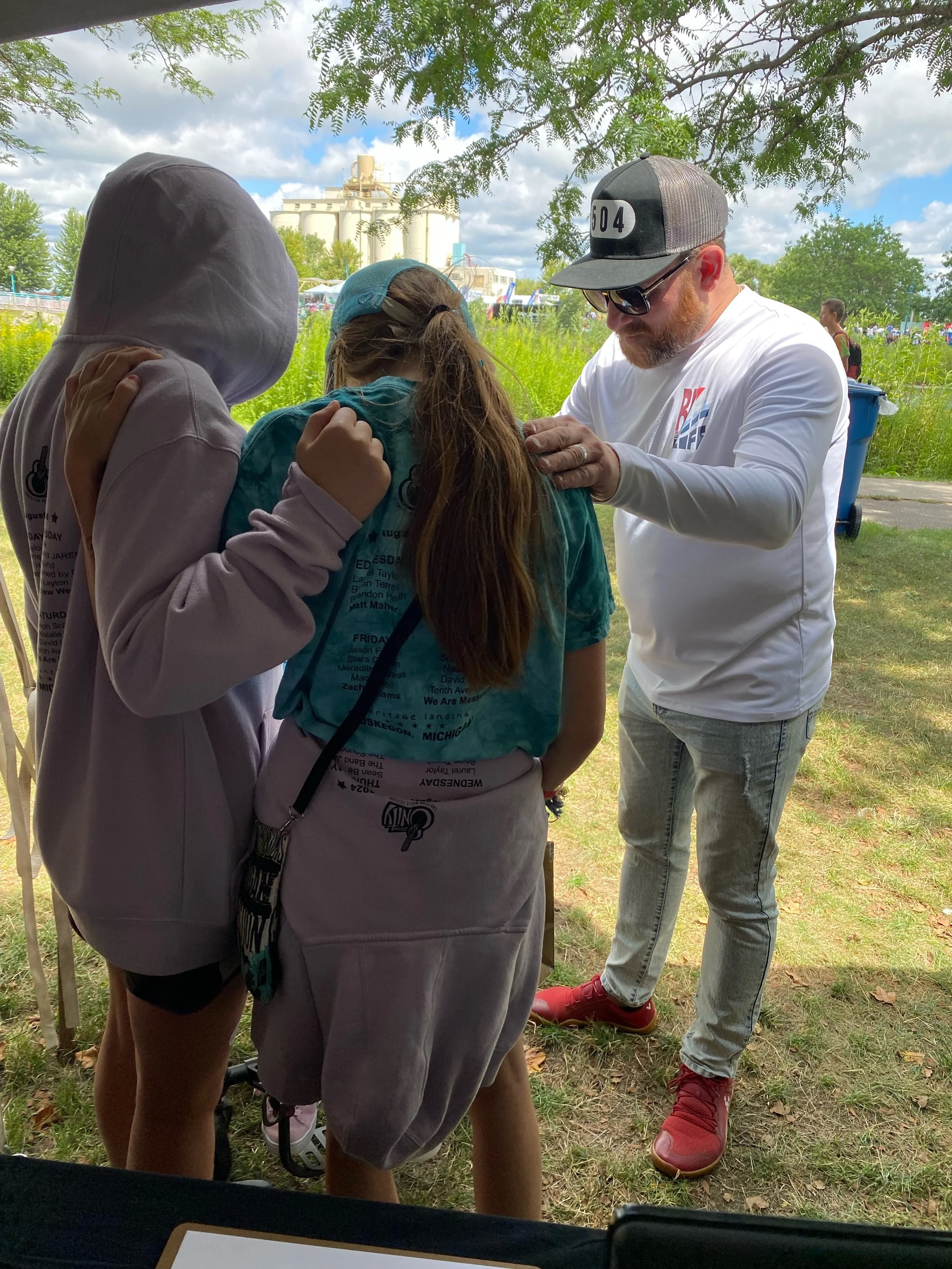 A man with a beard, wearing a black cap, sunglasses, and a white long-sleeve shirt, is praying at an outdoor event. They are standing on grass with trees and an industrial building or silo in the background.