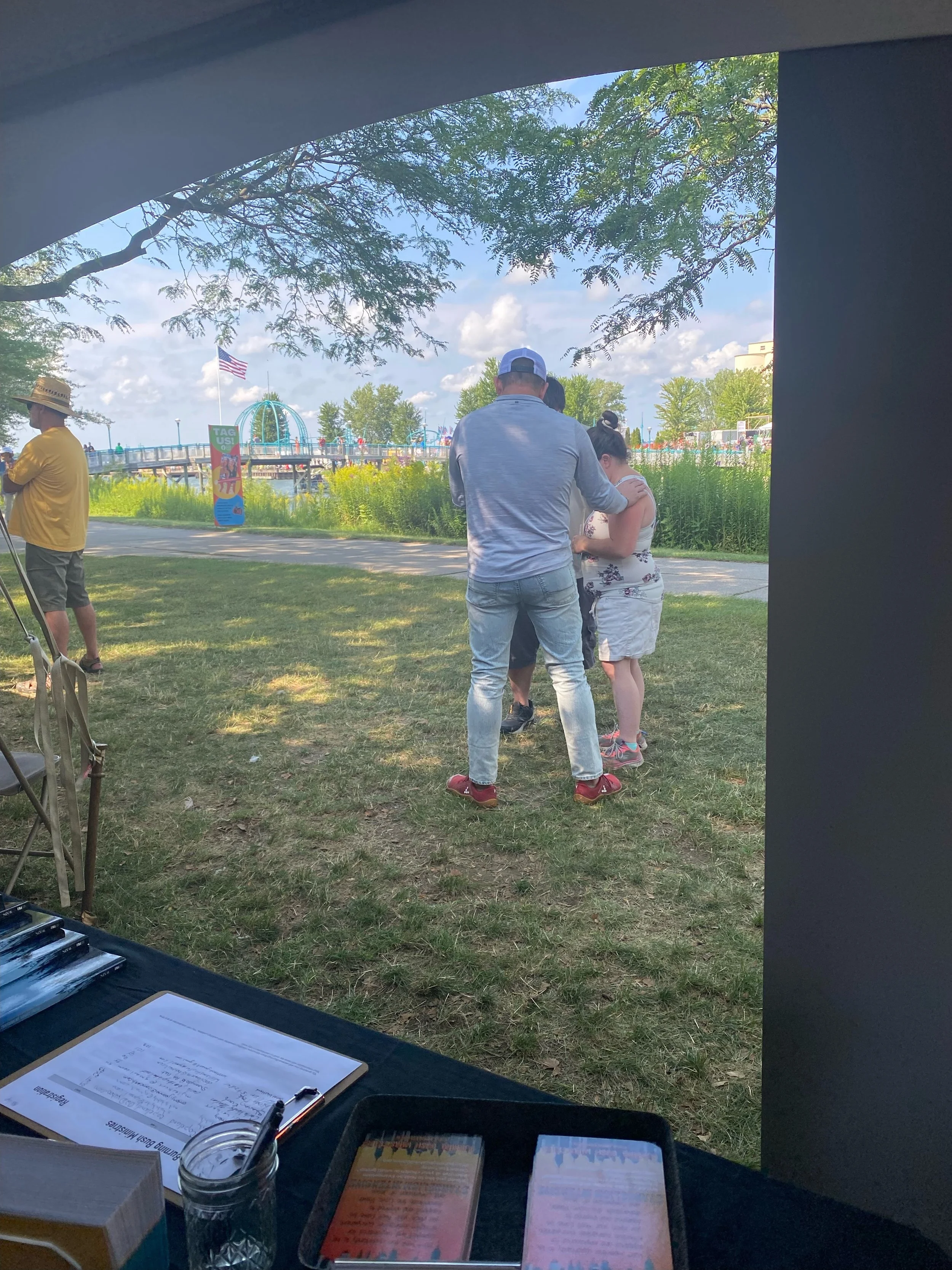 People standing outdoors praying, with a fence, trees, and a partly cloudy sky in the background.