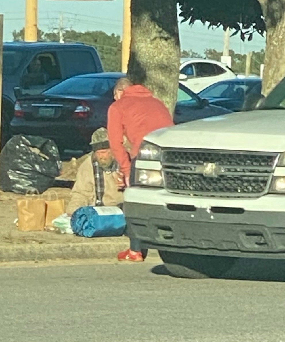 A person in a red jacket and red shoes leaning over a tree, talking to a man sitting on the ground with camping gear and bags around him, near parked cars.