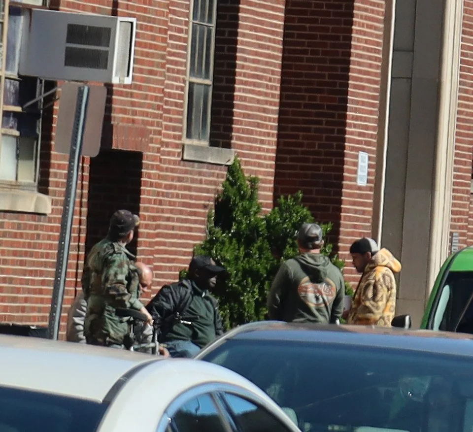 Four men gathered outside a brick building, some wearing hoodies and camouflage attire, near a parked car and a green vehicle, praying.
