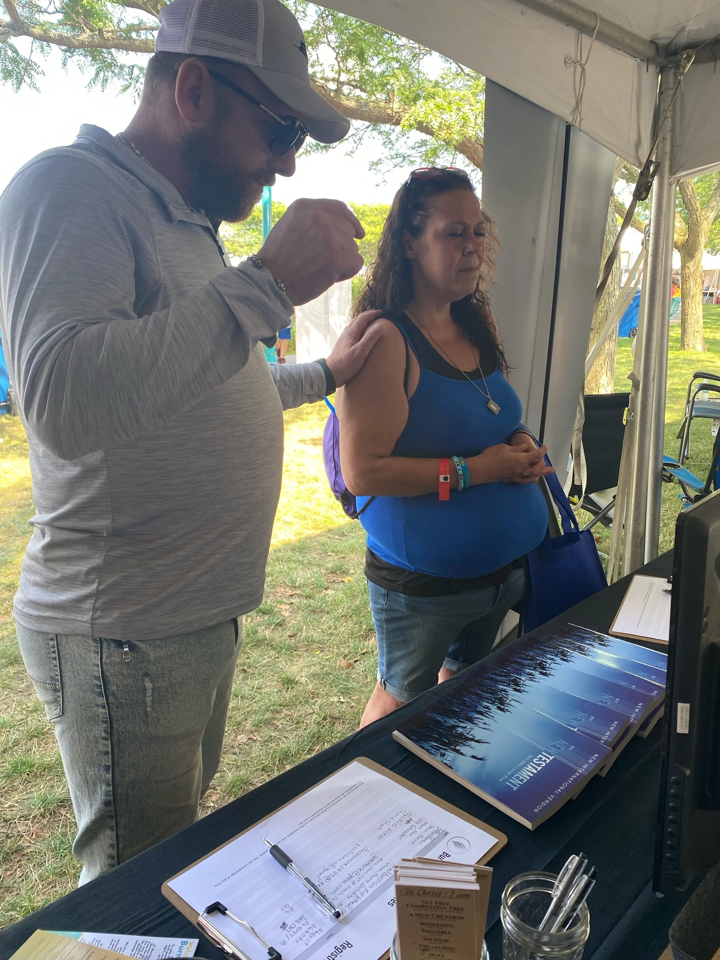 A man with a beard, sunglasses, and a hat prays for a woman as they stand near a table at an outdoor event. The woman has curly hair, is wearing a blue tank top, and looks at something on the table.