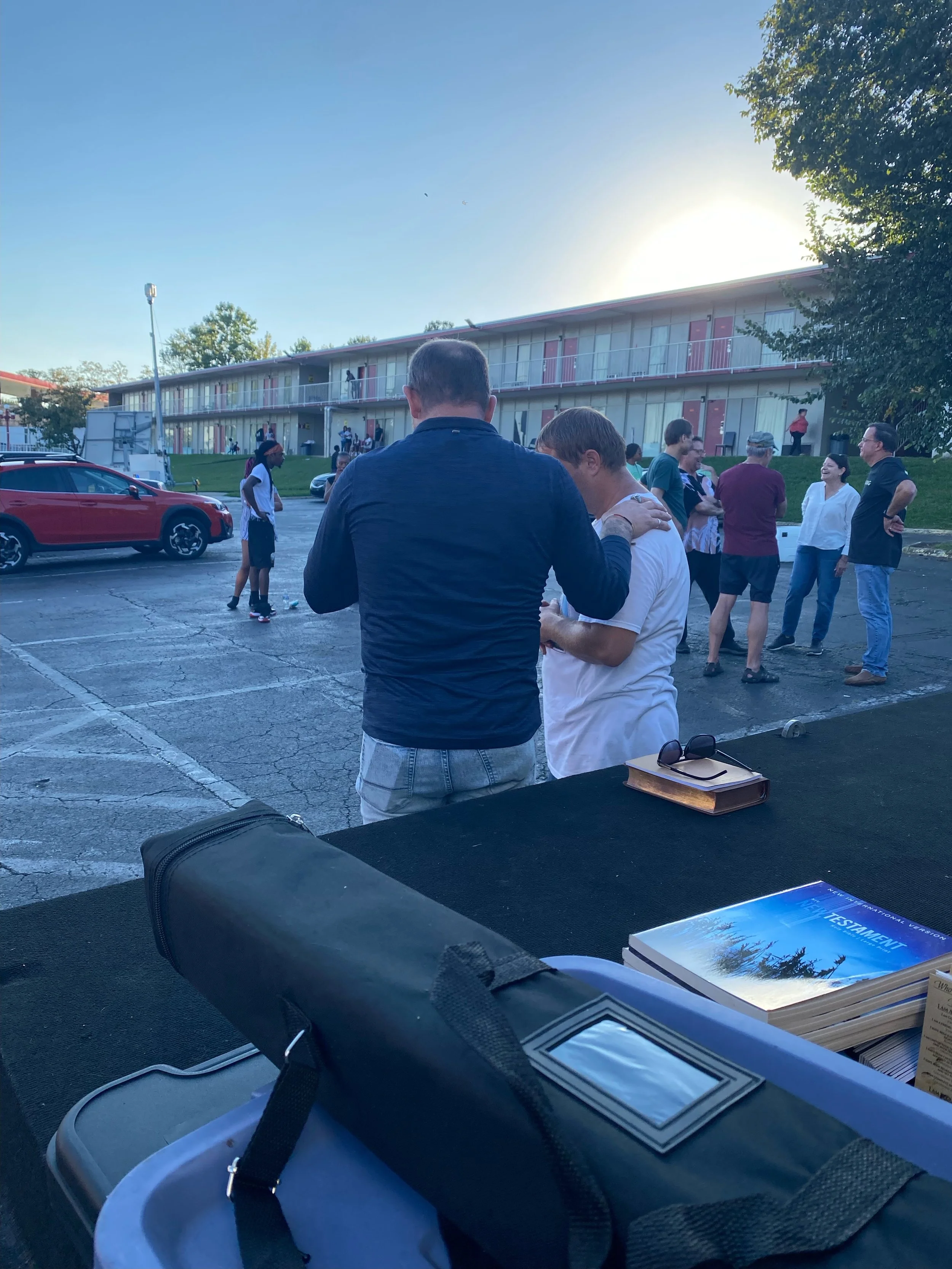 People gathered outdoors near a building with motel-style rooms, some engaging in prayer, with books and sunglasses on a table in the foreground, during sunset.