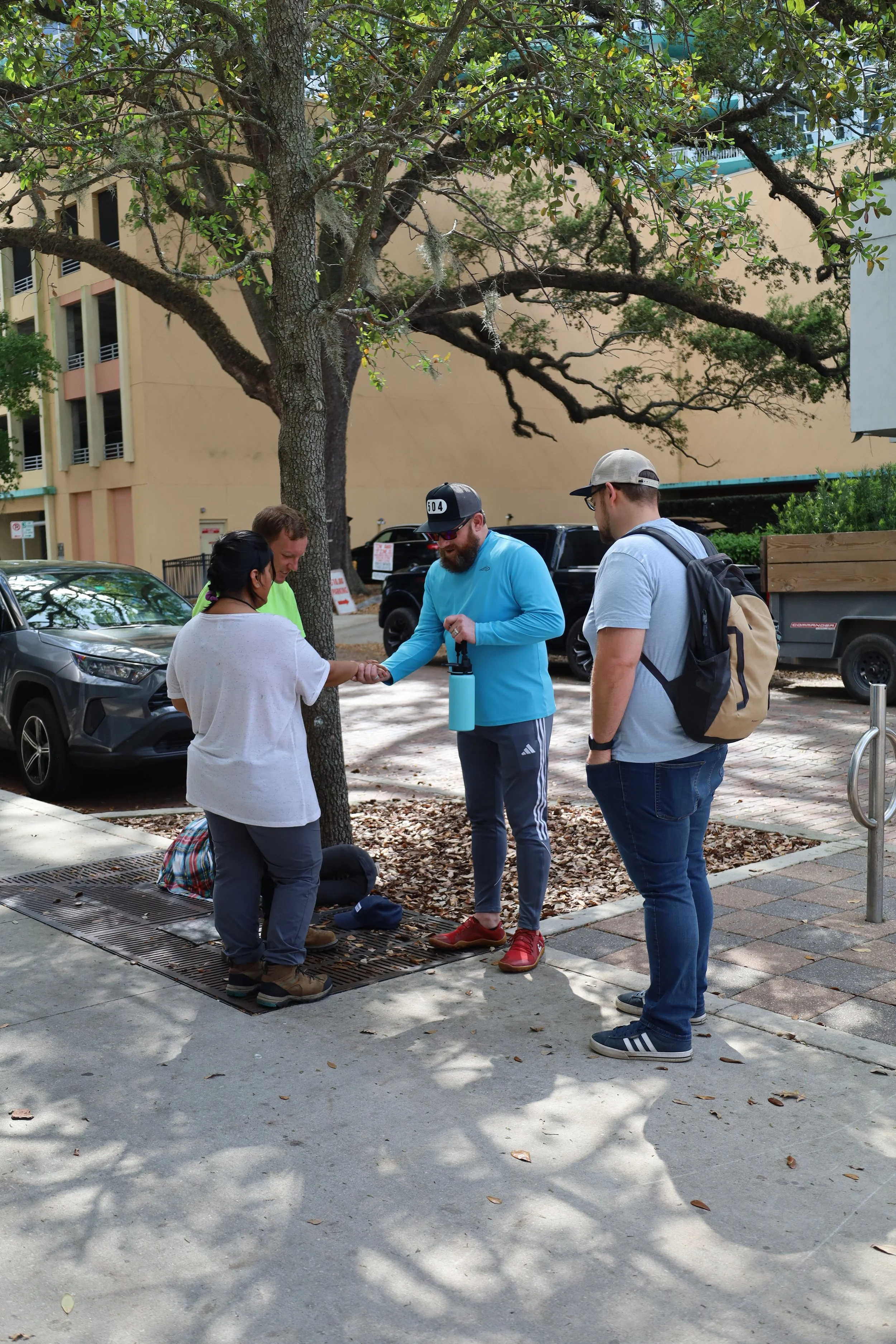 A group of four people standing on a sidewalk next to a tree, praying, with one person holding a water bottle. Three are standing, one appears to be kneeling or sitting on the ground, surrounded by parked cars and a building in the background.