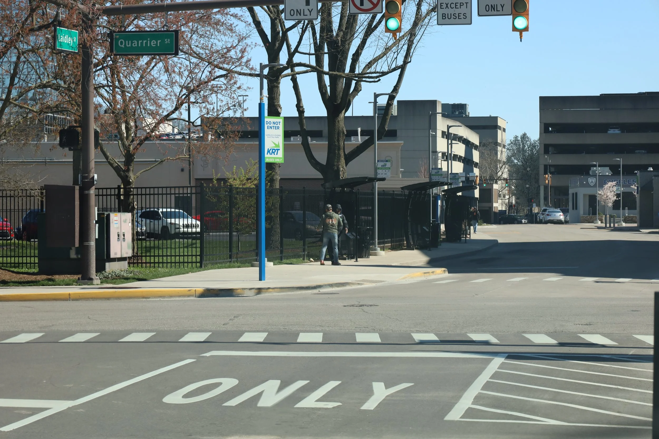 A city street scene with a crosswalk, traffic lights, and a bus stop. There are a few people waiting, surrounded by trees, parking lot, and buildings in the background.