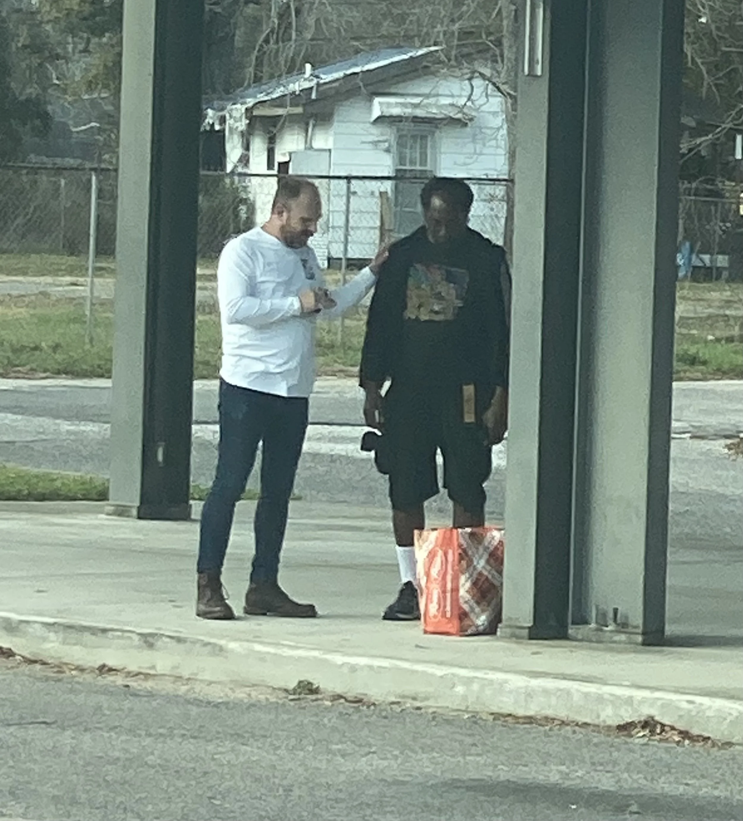 Two men standing at a bus stop praying; one dressed in a white shirt and jeans, the other in a black hoodie and shorts, holding a book and a bag, with a shopping bag on the ground nearby.