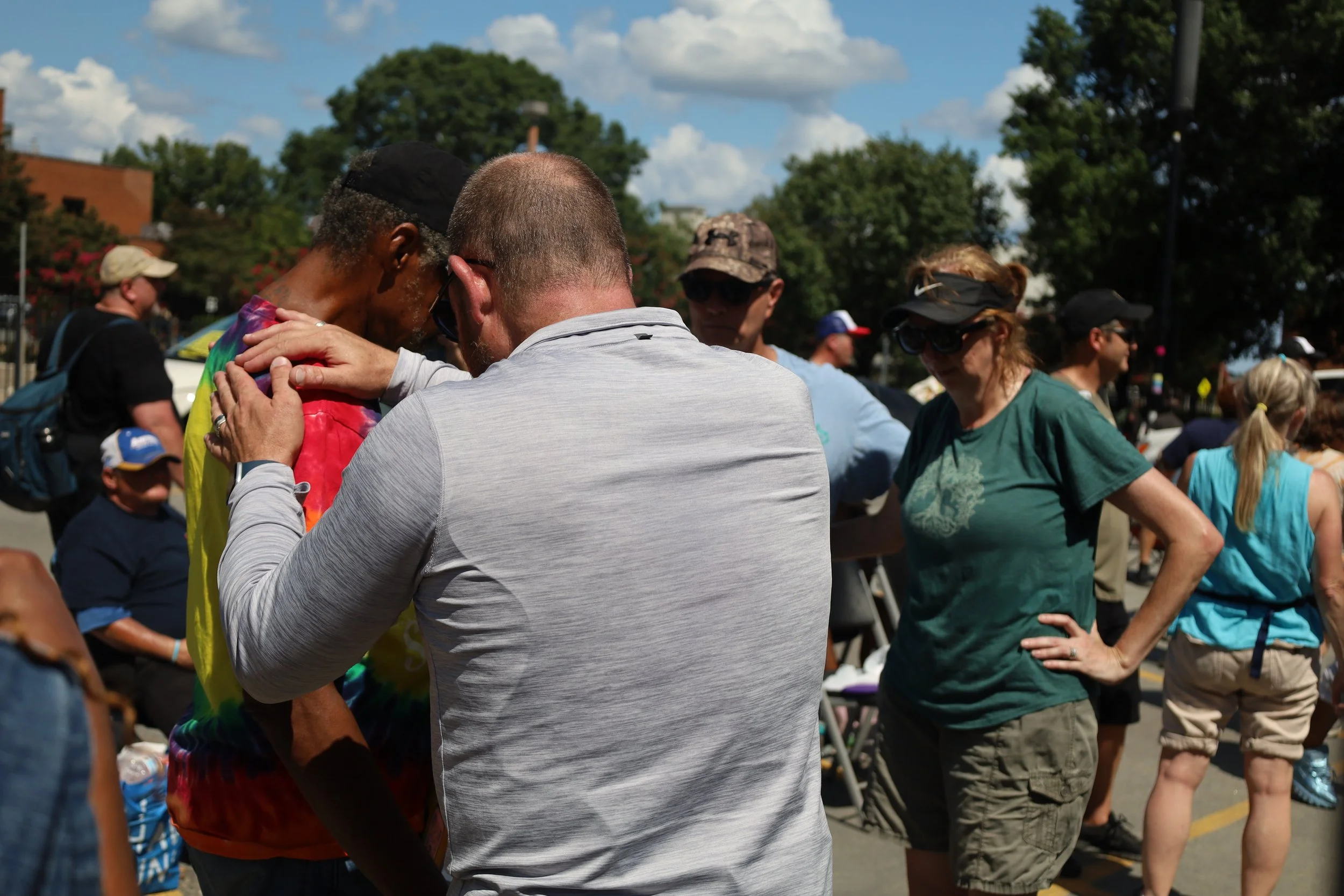 People praying outdoors in a crowd on a sunny day with trees in the background.