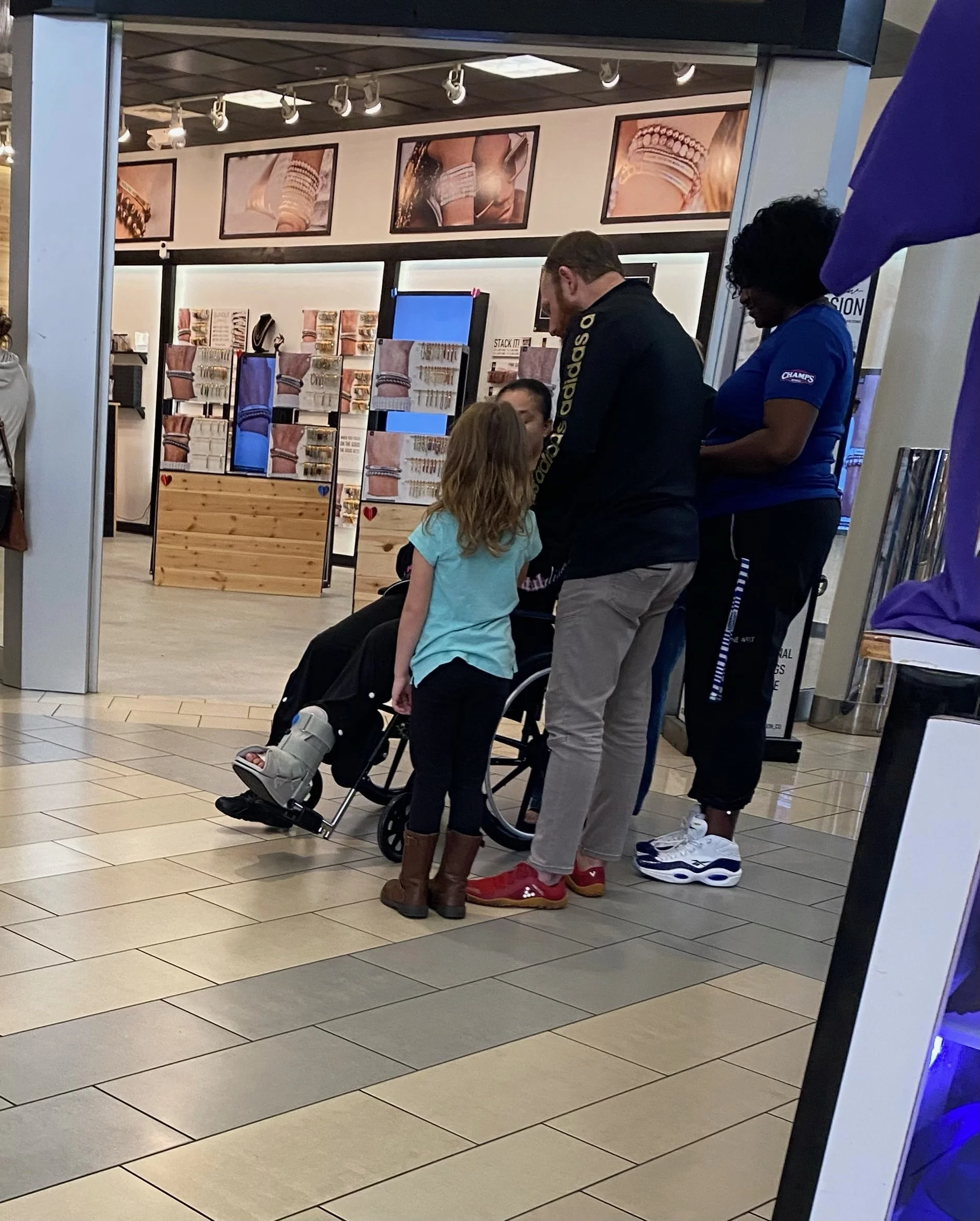 A man, a woman, a young girl, and a person in a wheelchair, are gathered in front of a jewelry store inside a shopping mall praying together. The store displays jewelry and accessories in the background.
