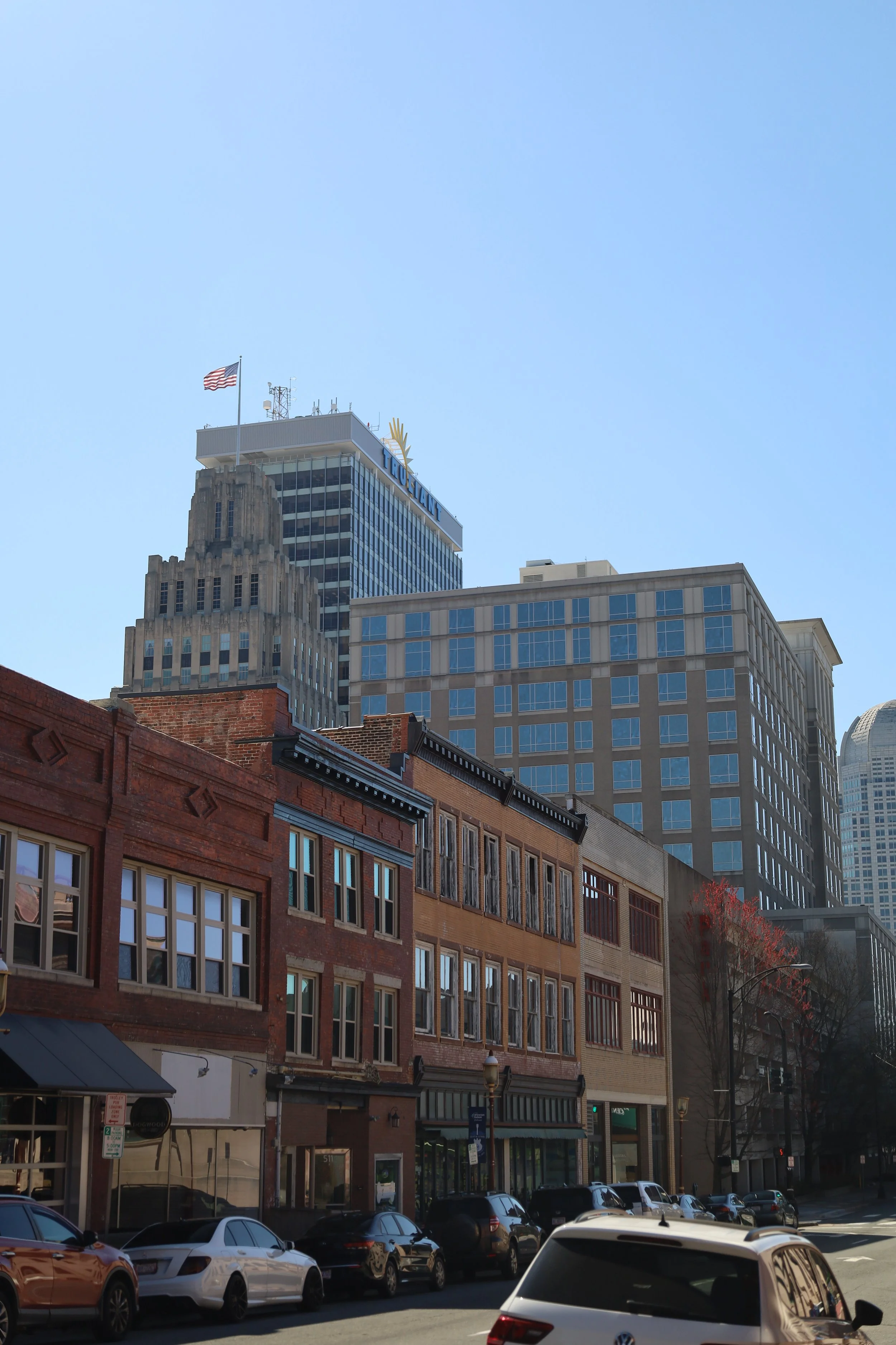 Downtown city scene featuring a row of older brick buildings with retail storefronts, cars parked along the street, and taller modern skyscrapers in the background under a clear blue sky.