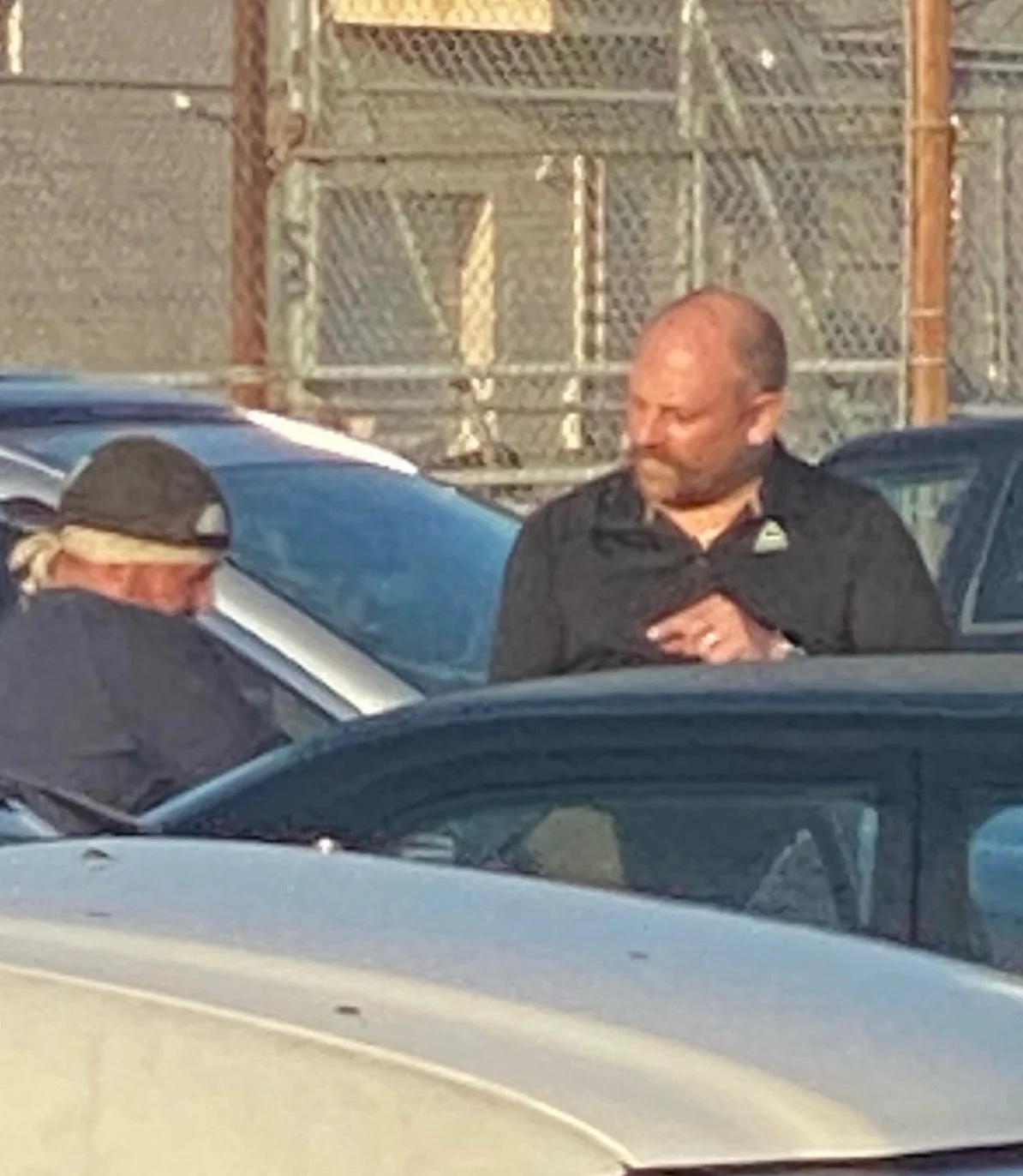 A man and a woman engage in conversation in a parking lot, with several cars and a chain-link fence in the background.