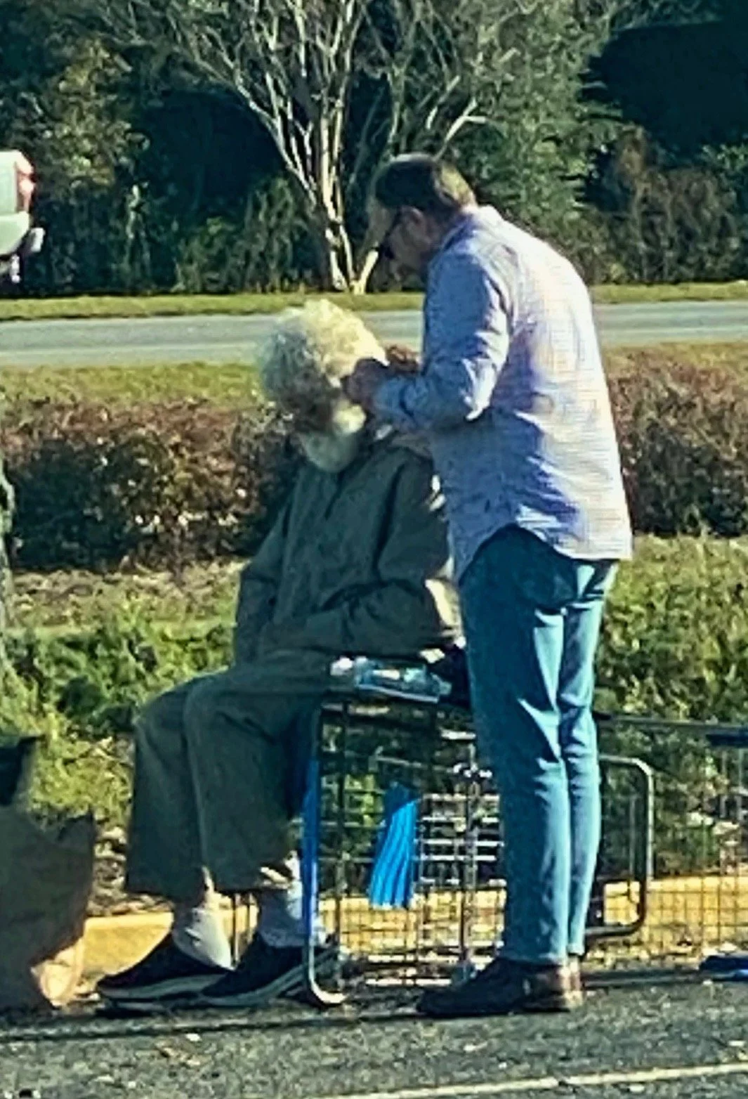 A person in a brown jacket sitting on a shopping cart, and another person in a light-colored jacket and jeans standing next to them, praying outdoors with greenery in the background.