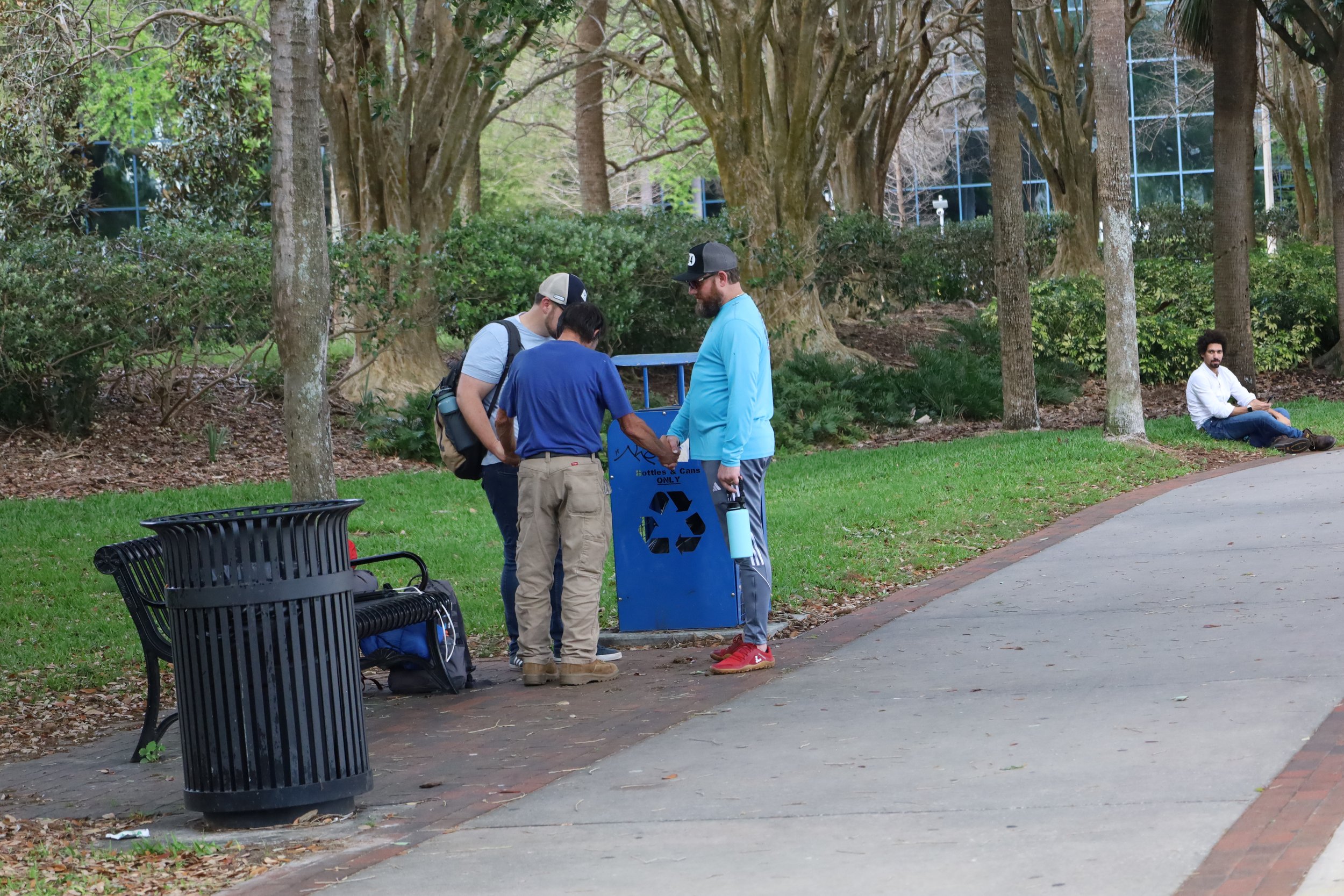 Three people in a park gathered around a recycling bin with a biodegradable cup, the three are praying. Another man sitting alone on a bench in the background.