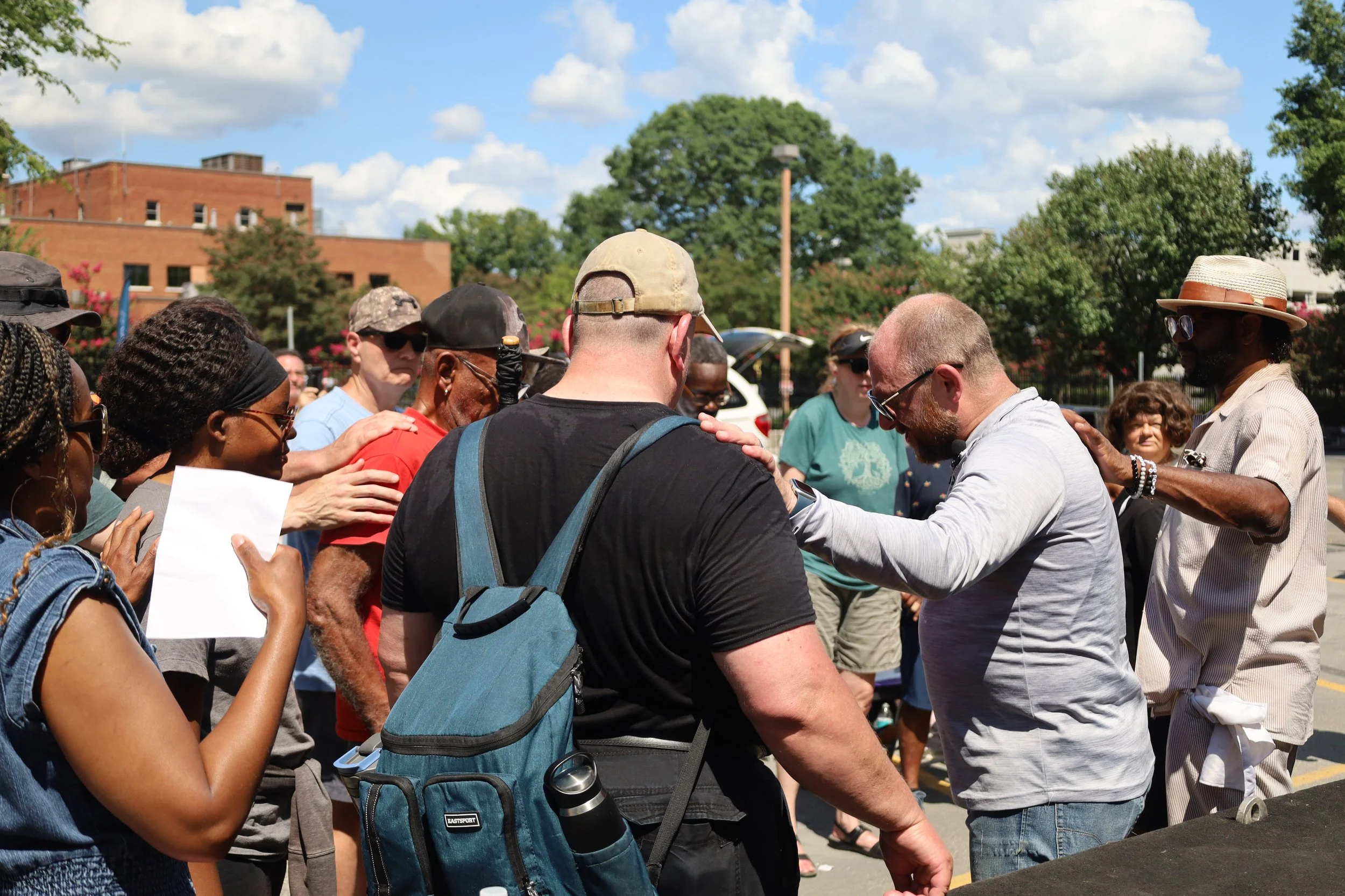 A group of diverse people standing outdoors in a circle, some with eyes closed, gathering around a man who is participating in a prayer on a sunny day with trees and buildings in the background.