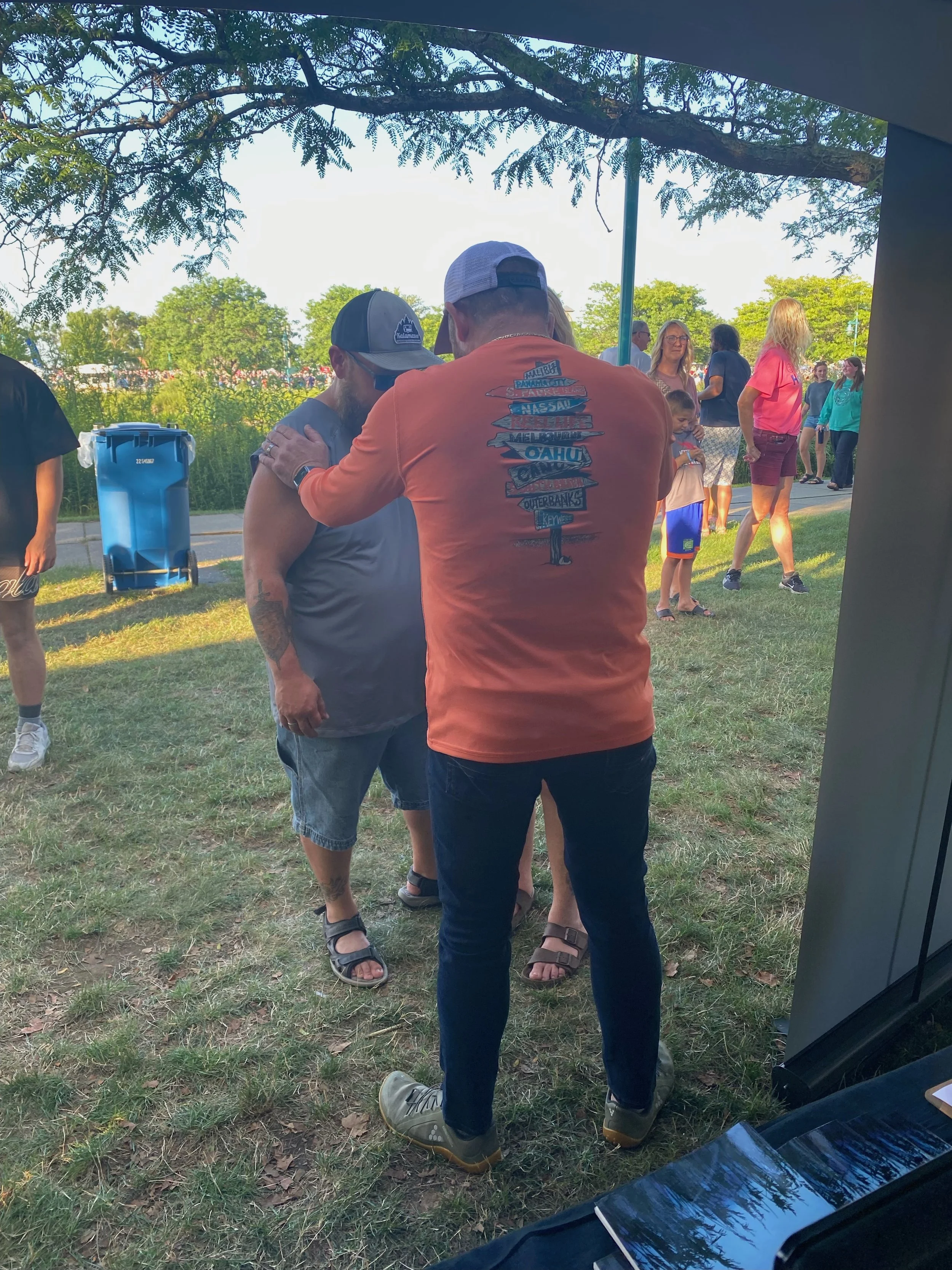 Two men are outdoors, one in a gray tank top and the other in an orange t-shirt, with the orange shirt having a design of a signpost with different place names. They are engaged in a close conversation. Several people, including children, are visible