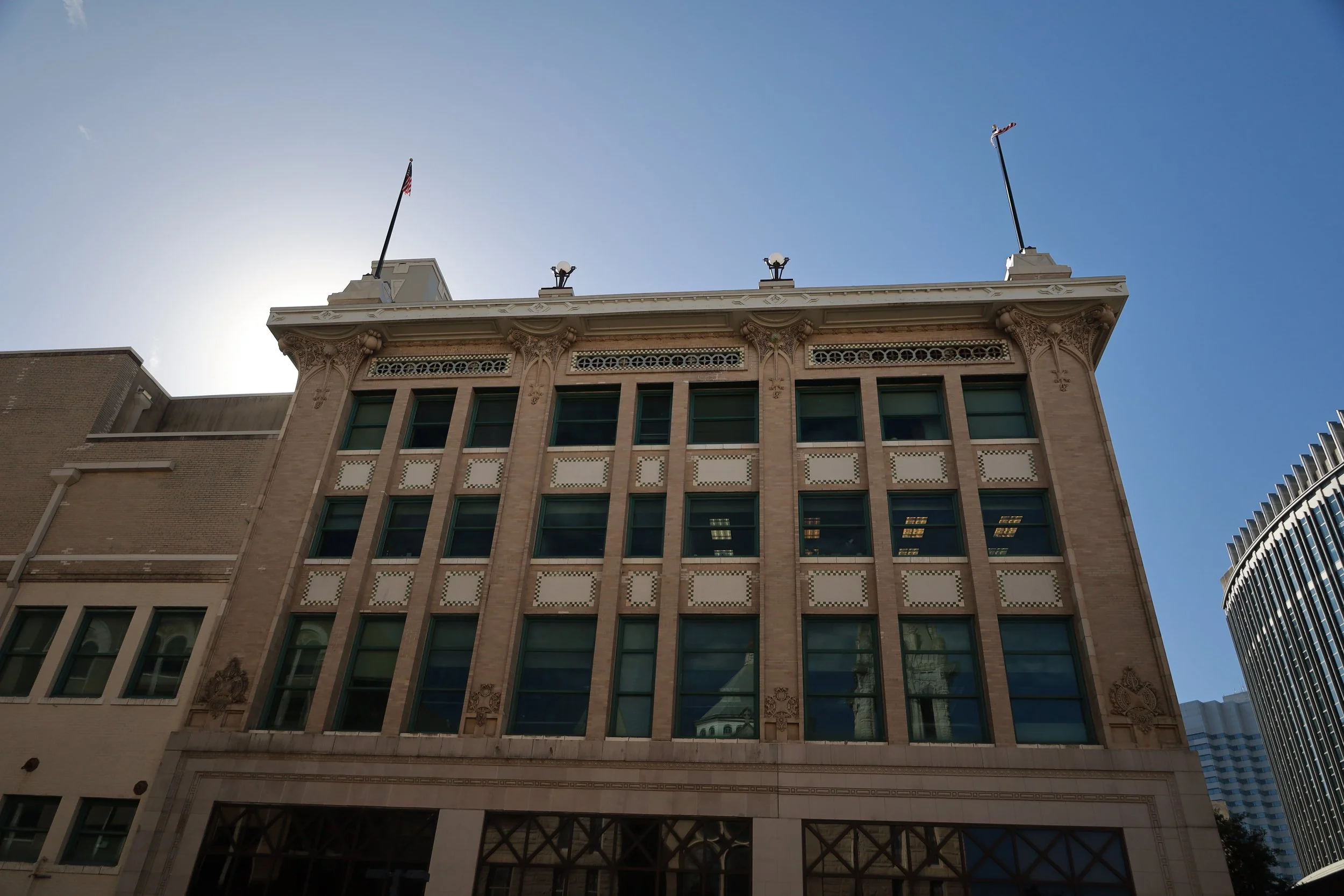 The facade of a historic multi-story building with decorative architectural details and flagpoles on the roof, set against a clear blue sky.