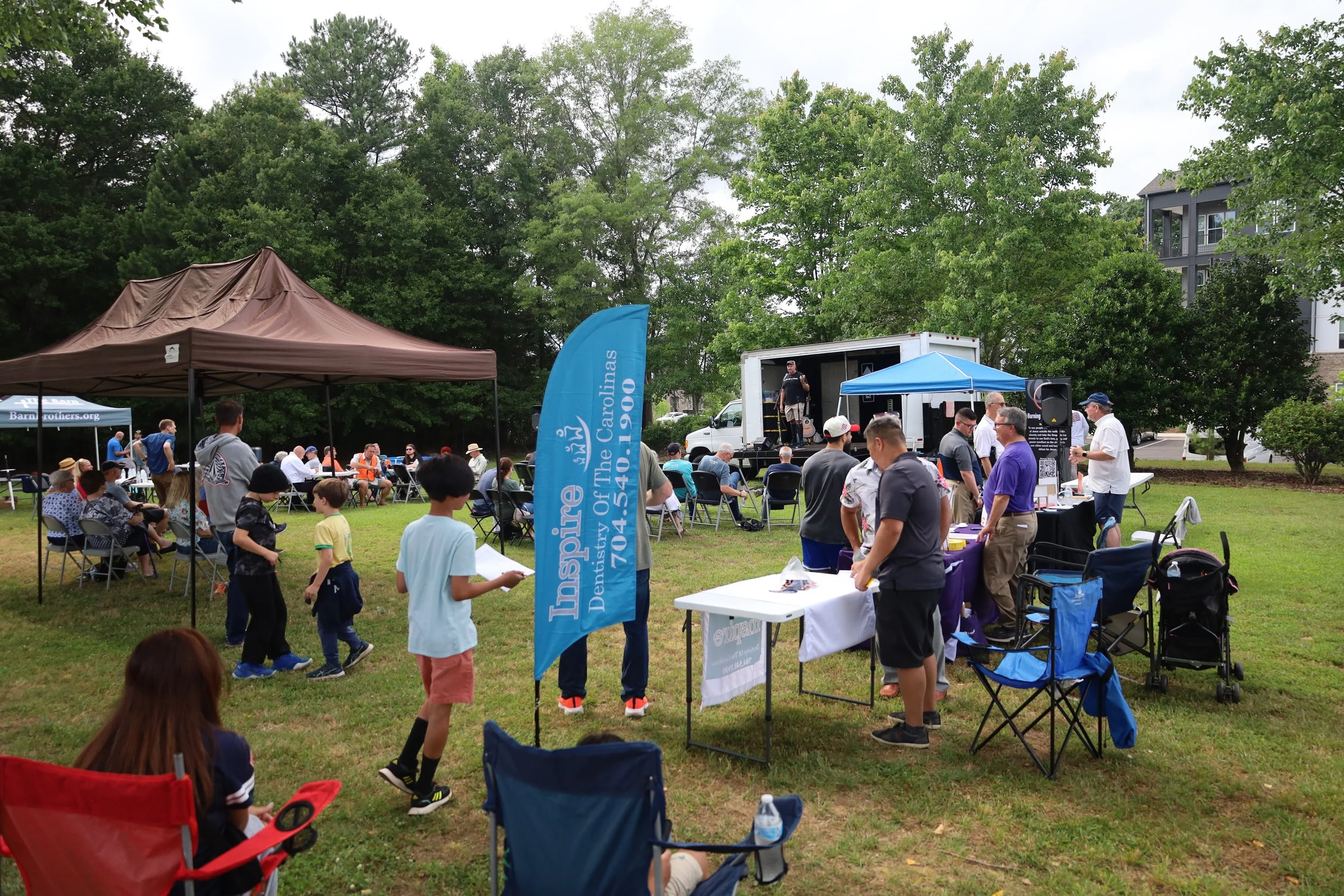 A community outdoor event with people gathered around tents, tables, and a stage in a park with green trees. Some people are sitting, some standing, and some are walking. There is a blue banner that reads 'Inspire of the Carolinas' and a man speaking