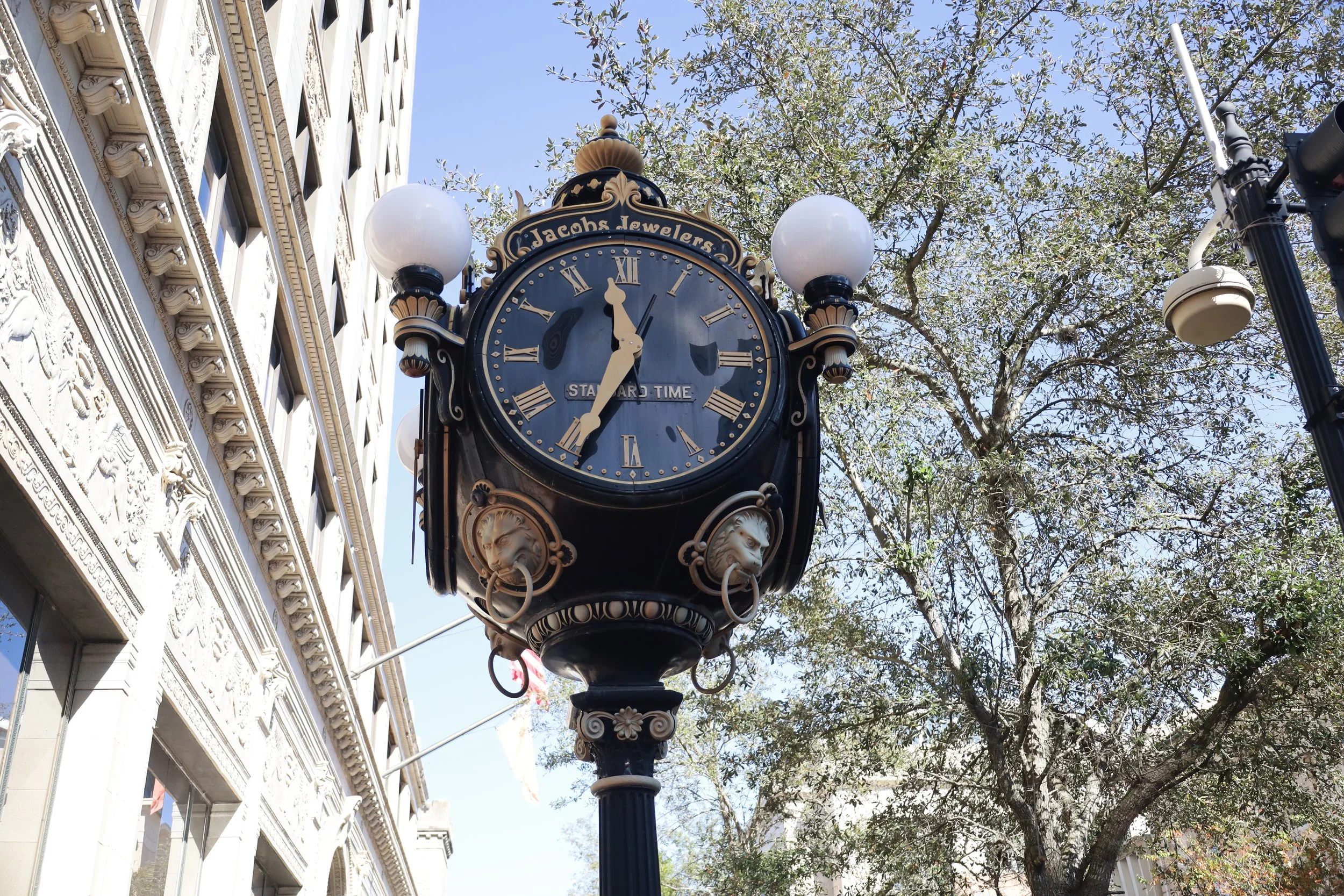 A vintage-style street clock with Roman numerals and decorative lion face details, displaying the time as 11:32, located outside near a building and trees.