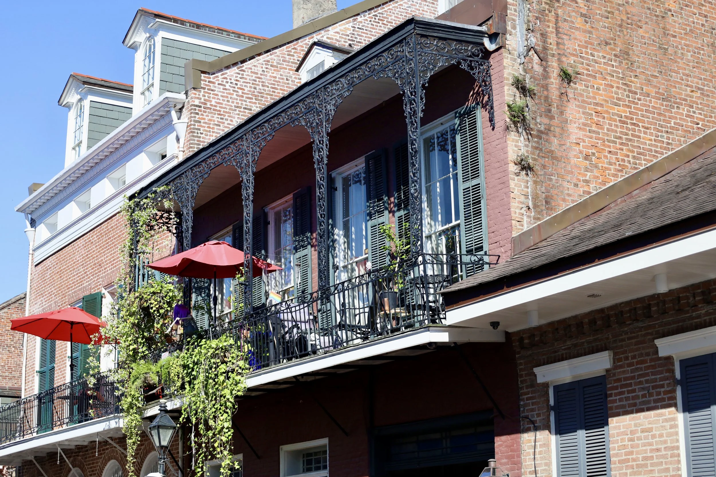 Brick apartment building with a balcony featuring black ornate metalwork, red umbrellas, and potted plants, under a clear blue sky.