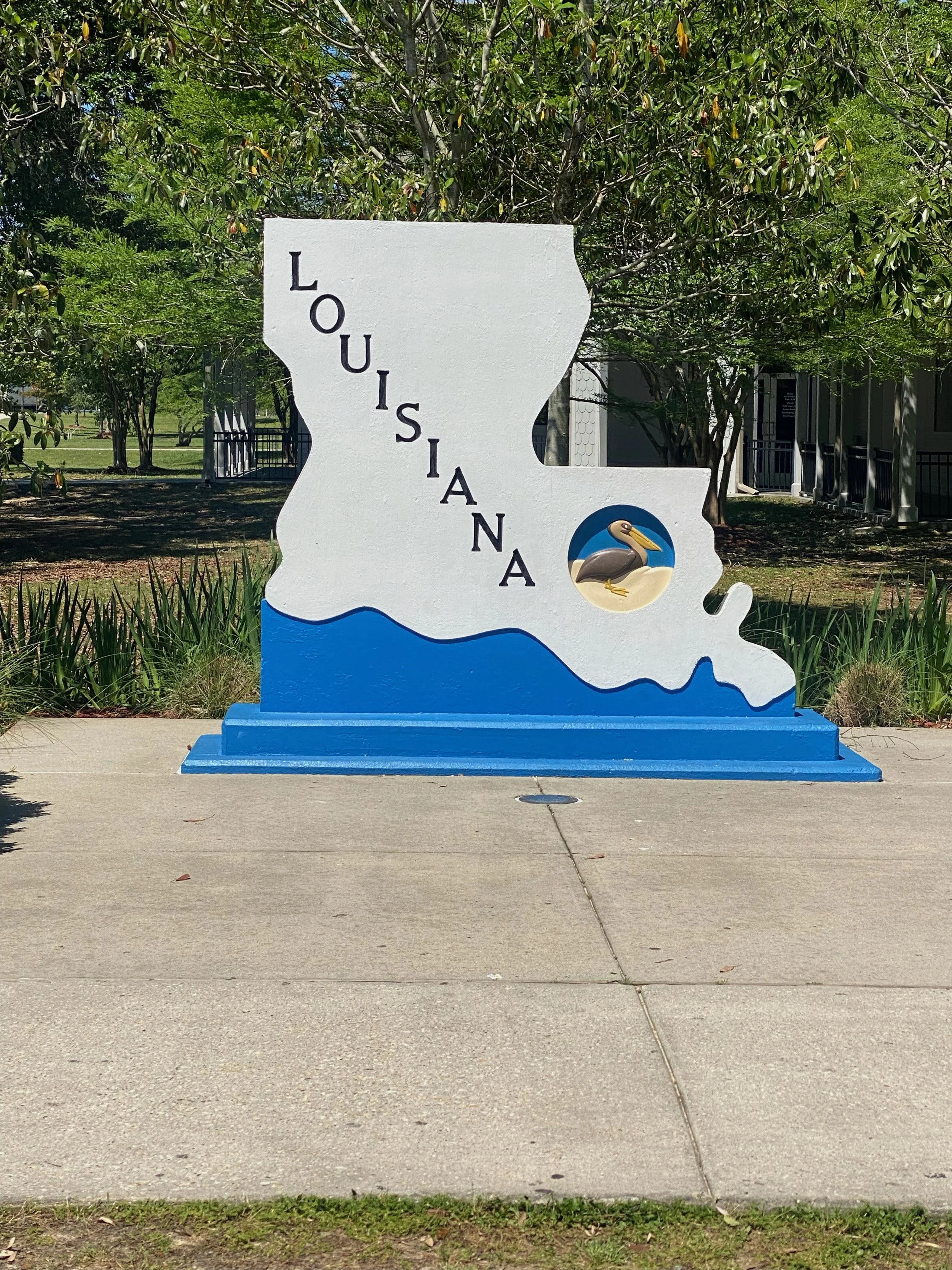 A decorative sign in the shape of Louisiana with the word 'Louisiana' written vertically across it, featuring a pelican illustration on the right side, and painted in white and blue colors. The sign is placed outdoors on a sidewalk with trees and a b
