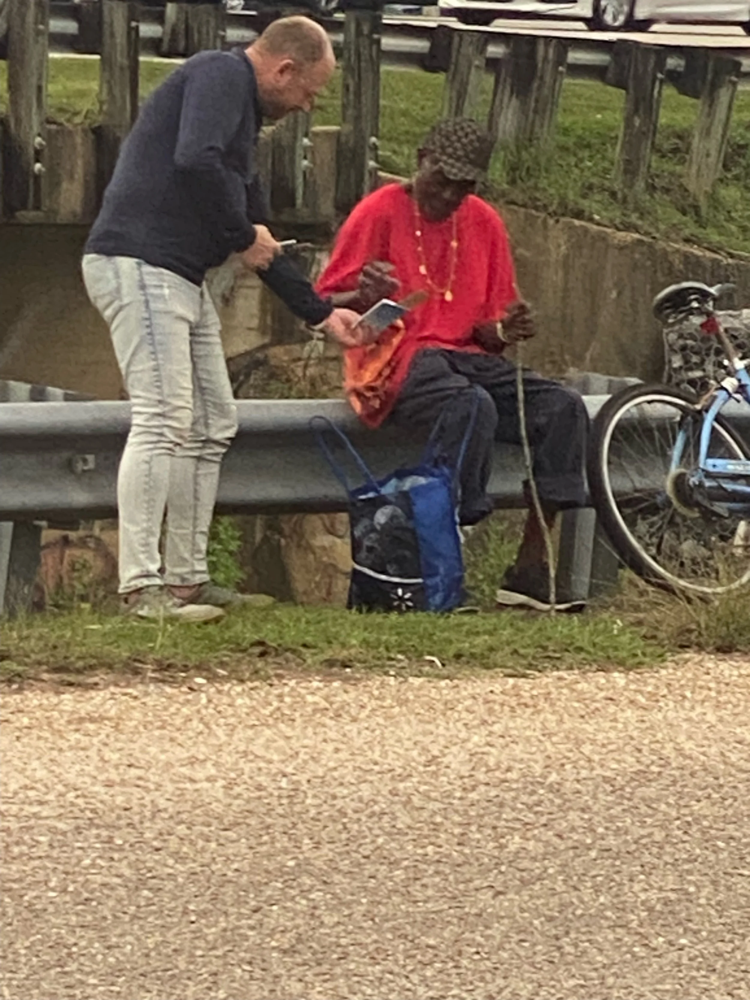 A man and a man sitting on a guardrail beside a creek. The man is dressed in bright red and black, wearing a hat and necklace. The man is dressed casually in light pants and a dark jacket. There is a bicycle leaning beside them.