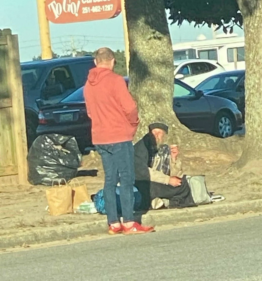 Two men are seen on a sidewalk near a street with parked cars and trees praying. One man, standing, wears a red hoodie and blue jeans. The other man, seated on the ground, wears a black hat, black jacket, and beige pants.