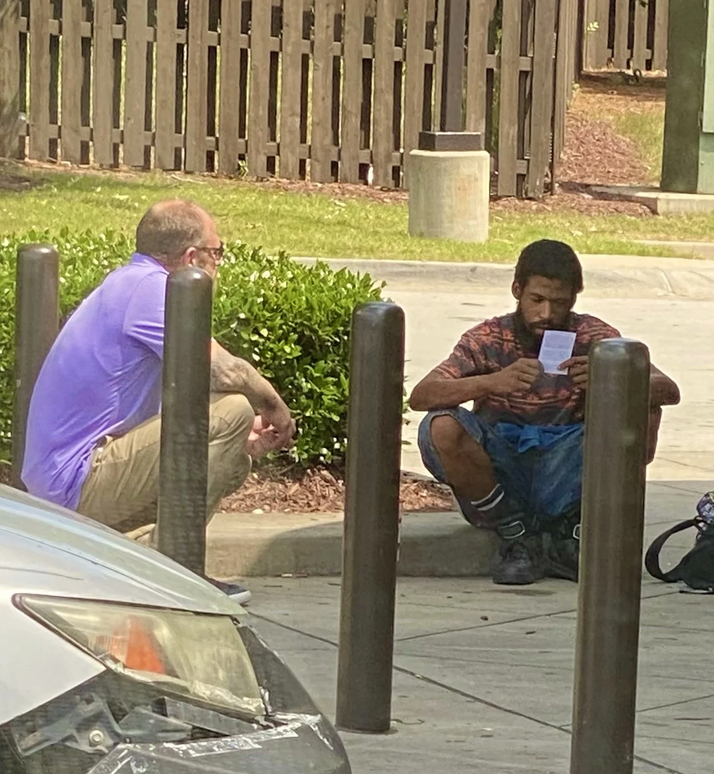 Two men sitting on the sidewalk behind protective posts, with one man reading something and the other man praying.
