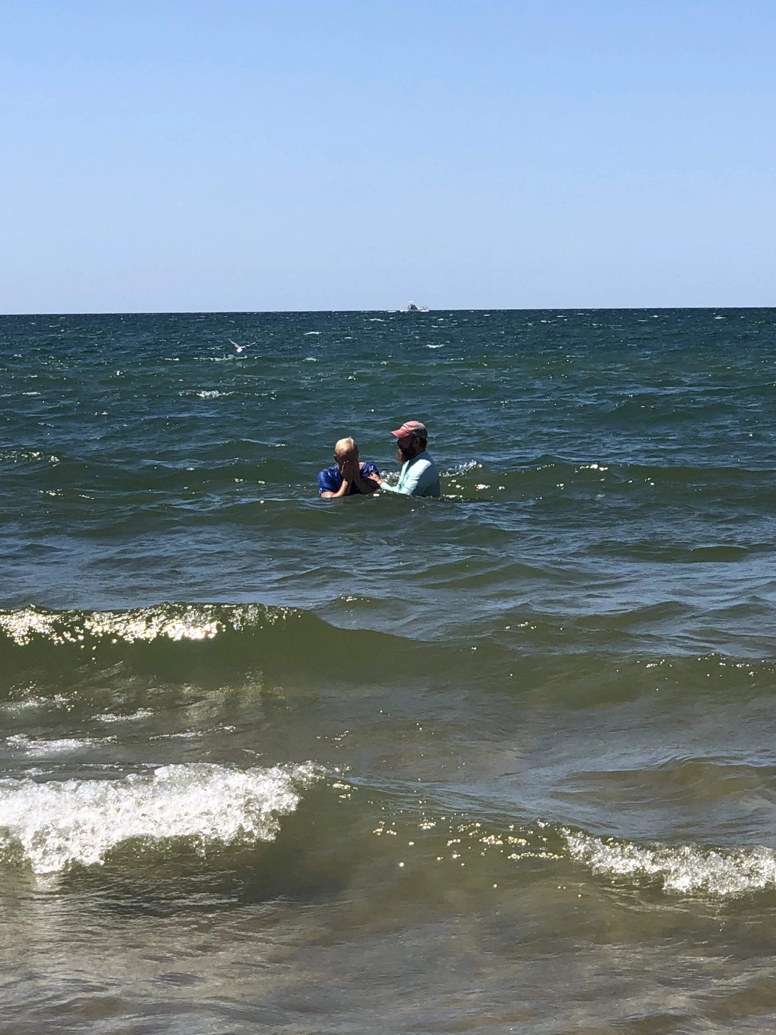 Two people standing in the lake for a water baptism, with a boat in the distance on a sunny day.