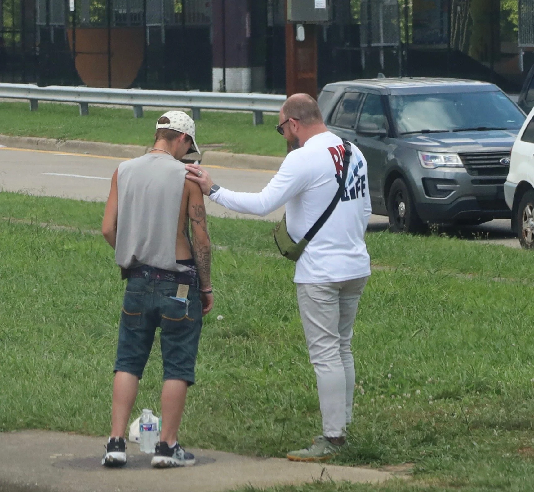 Two men standing on a grassy sidewalk; One man is praying with a hand on the others shoulder.