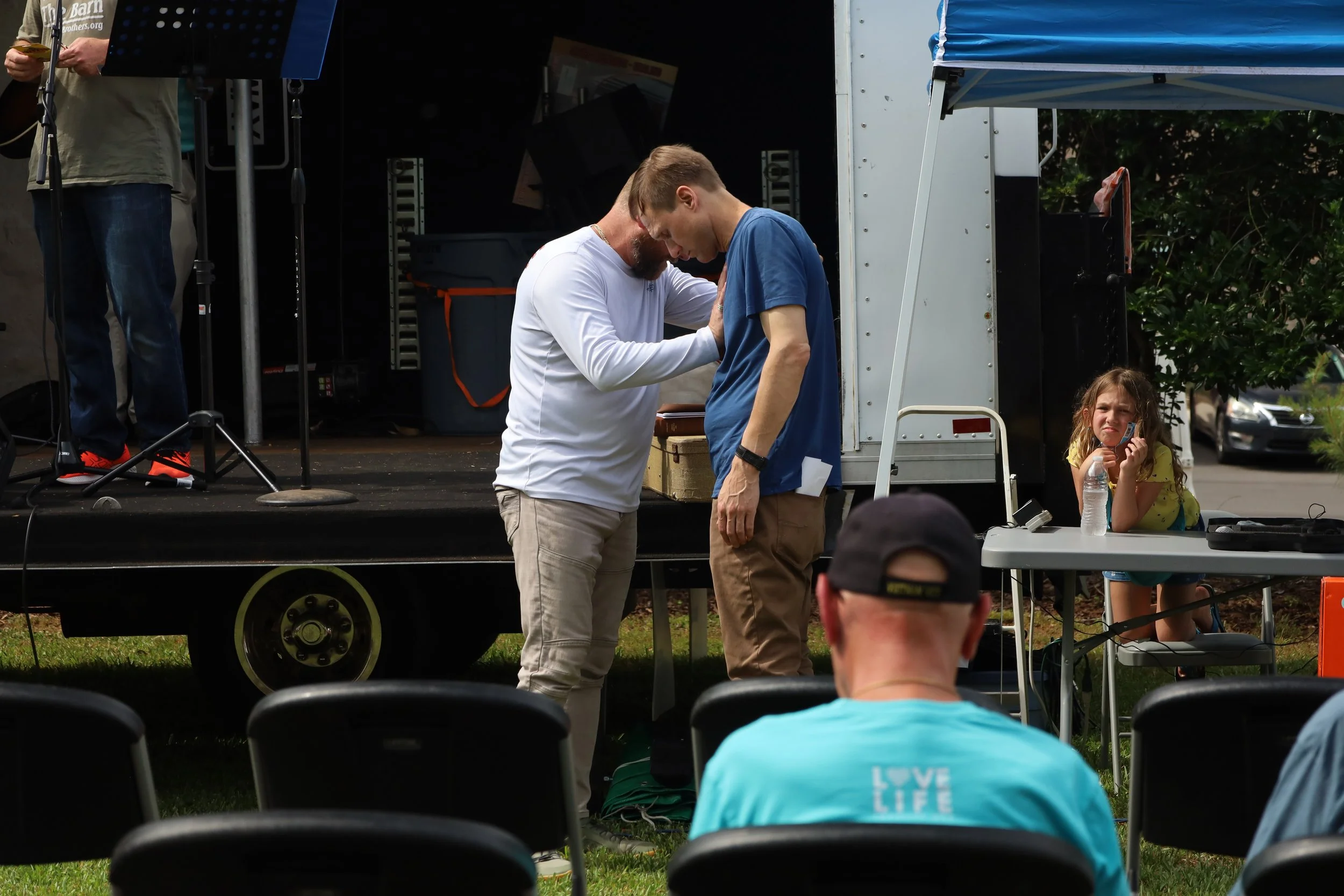 Two men are near a stage truck praying. A girl sitting at a table nearby talking on the phone and holding a water bottle. Several chairs and a person wearing a cap are in the foreground.