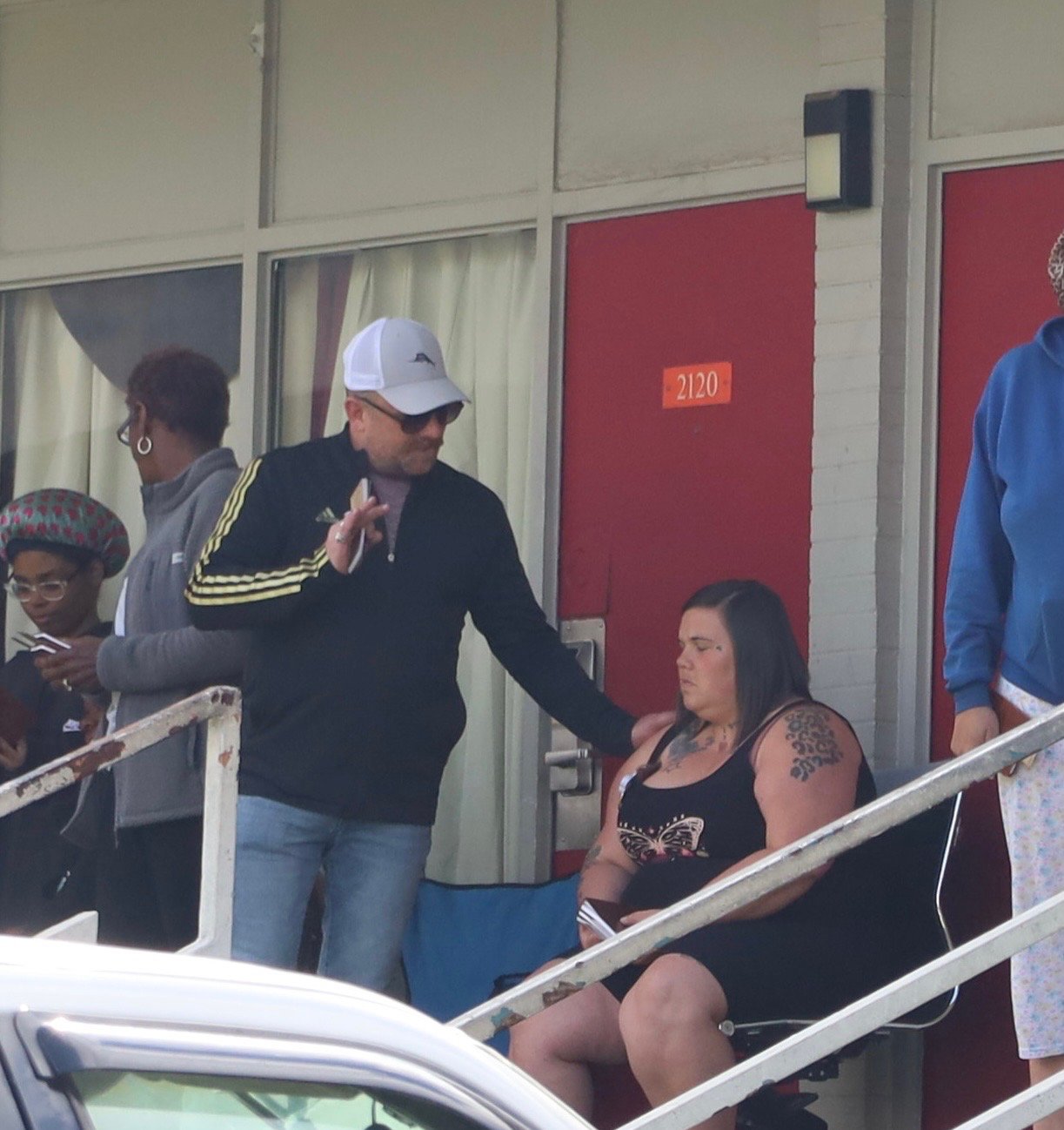 A woman with tattoos sitting in a chair outside a building while a man with sunglasses and a white cap places a hand on her shoulder and prays for her.