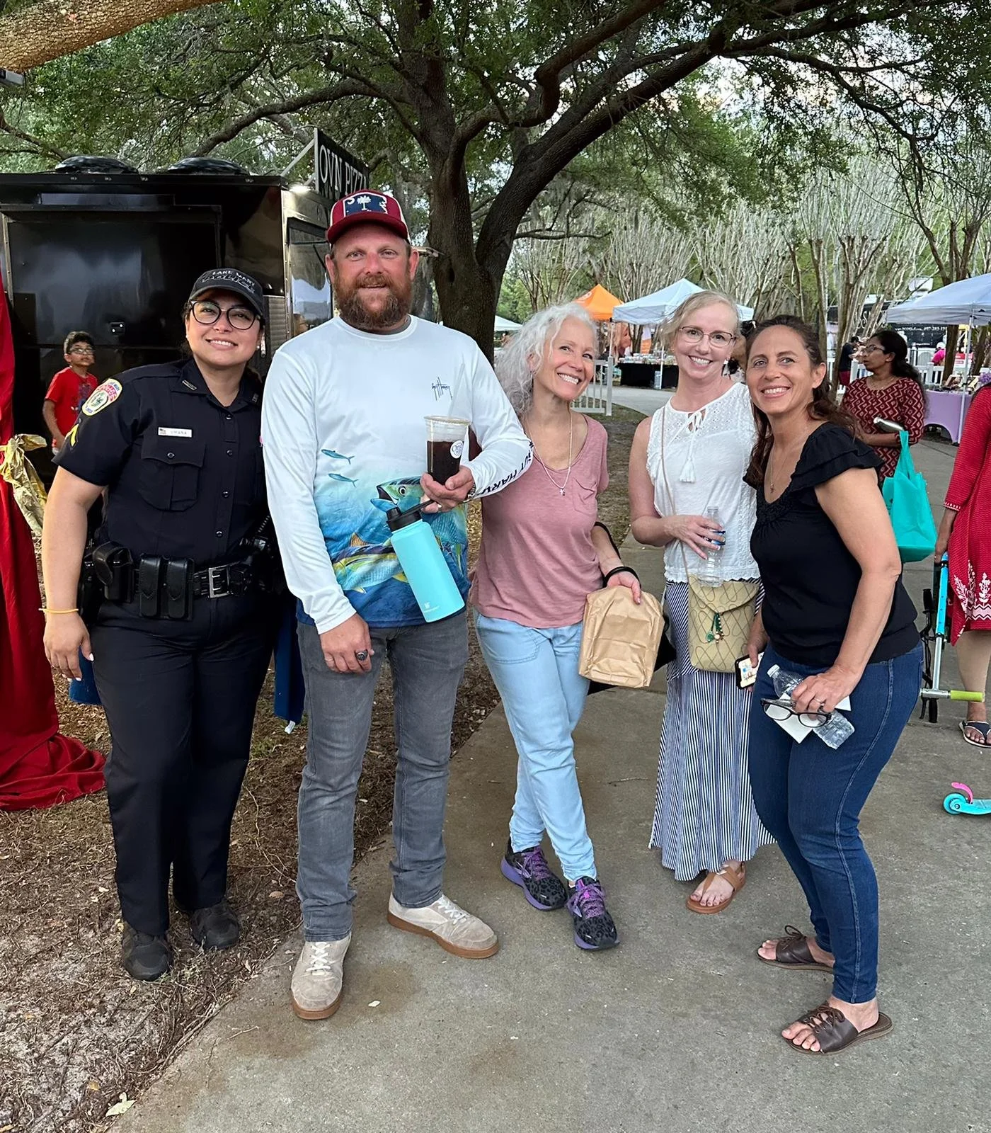 Group of five people, including a police officer, standing outdoors at a market or fair, smiling at the camera, with tents and trees in the background.