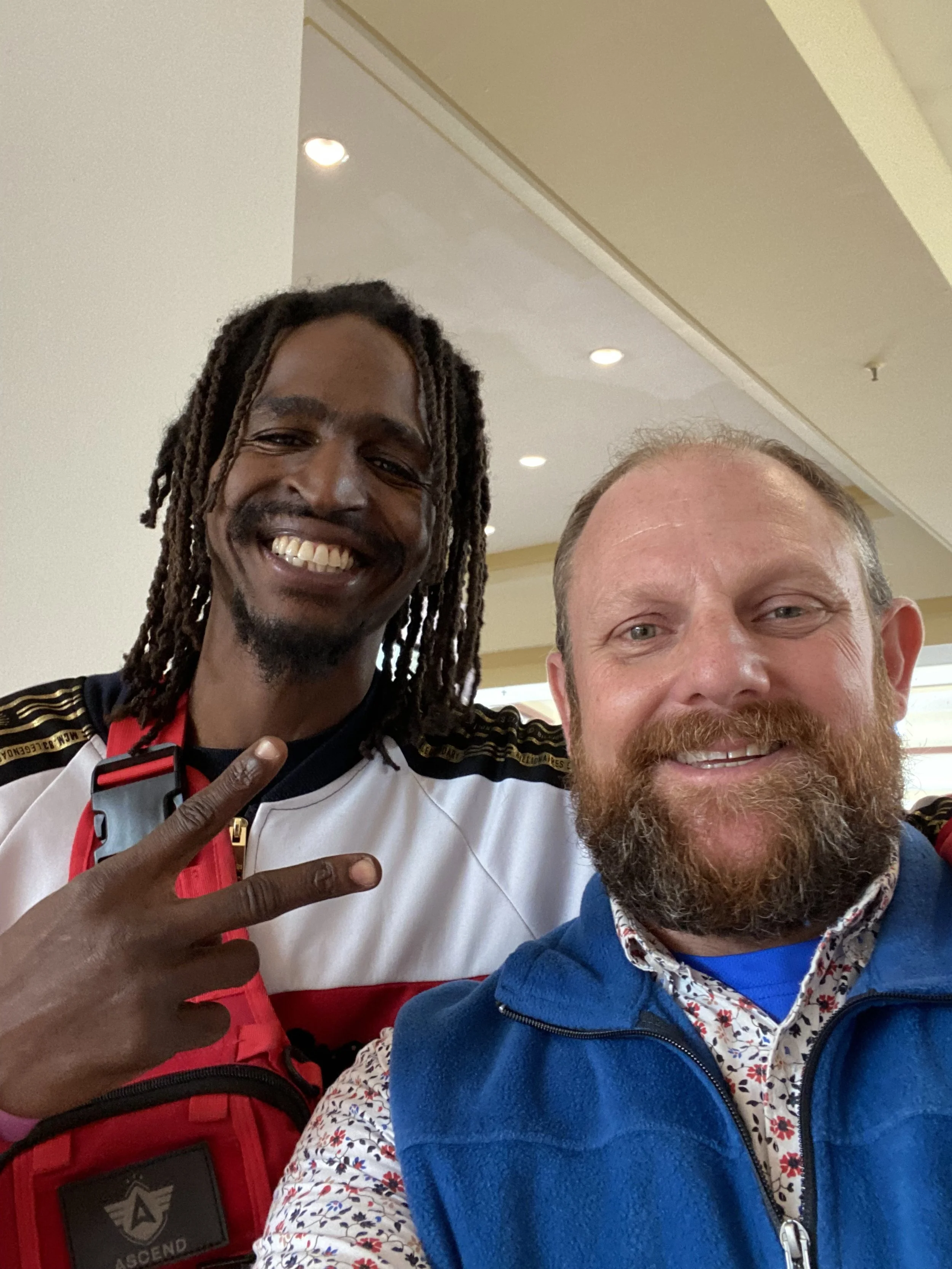 Two smiling men taking a selfie together in an indoor setting. The man on the left has long dreadlocks, dark skin, and is making a peace sign. He is wearing a white, black, and red jacket. The man on the right has light skin, a beard, and is wearing 