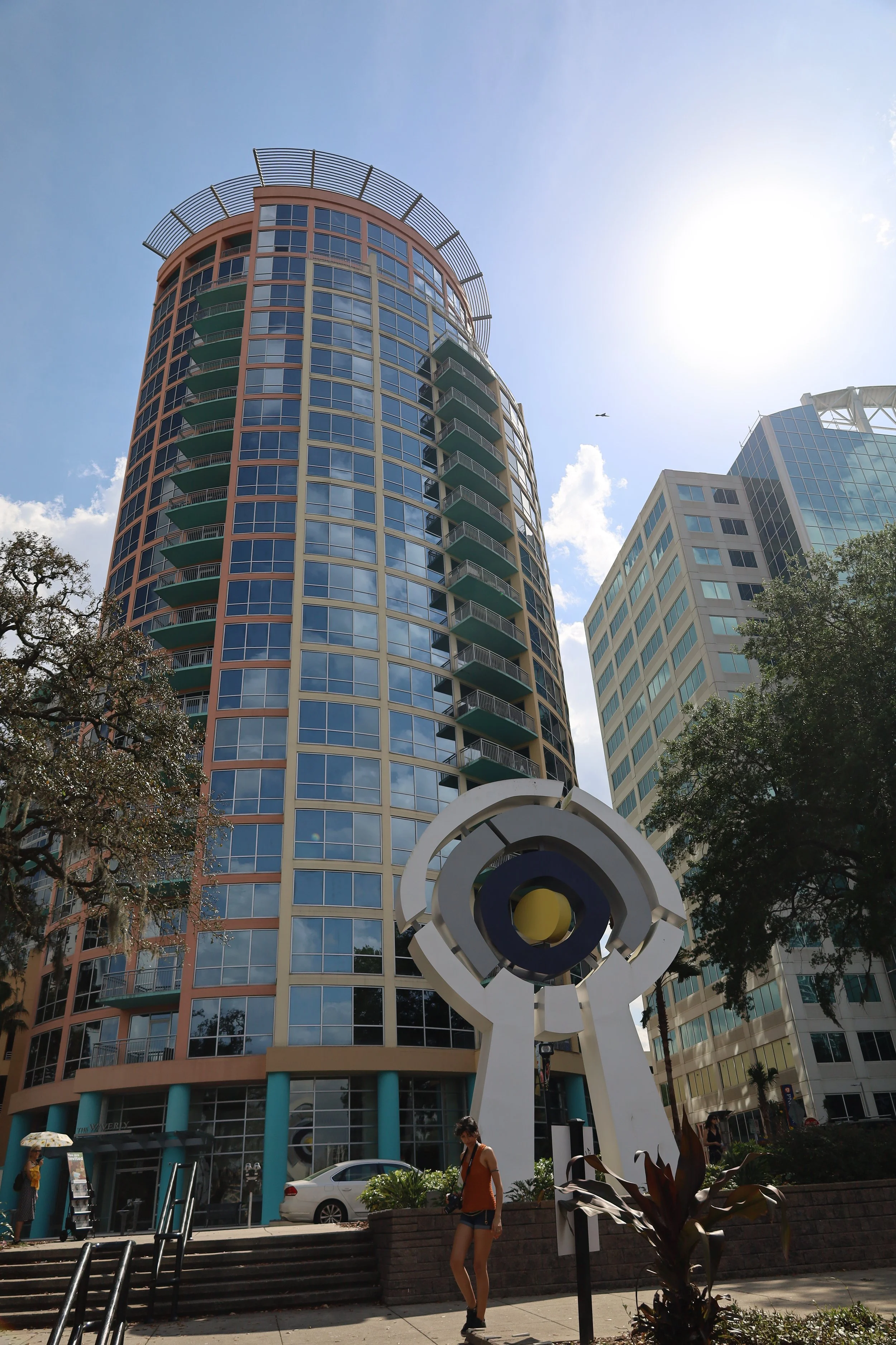 A tall, modern, circular glass apartment building with balconies on each floor, set against a bright blue sky with some clouds, and a large abstract sculpture in the foreground.