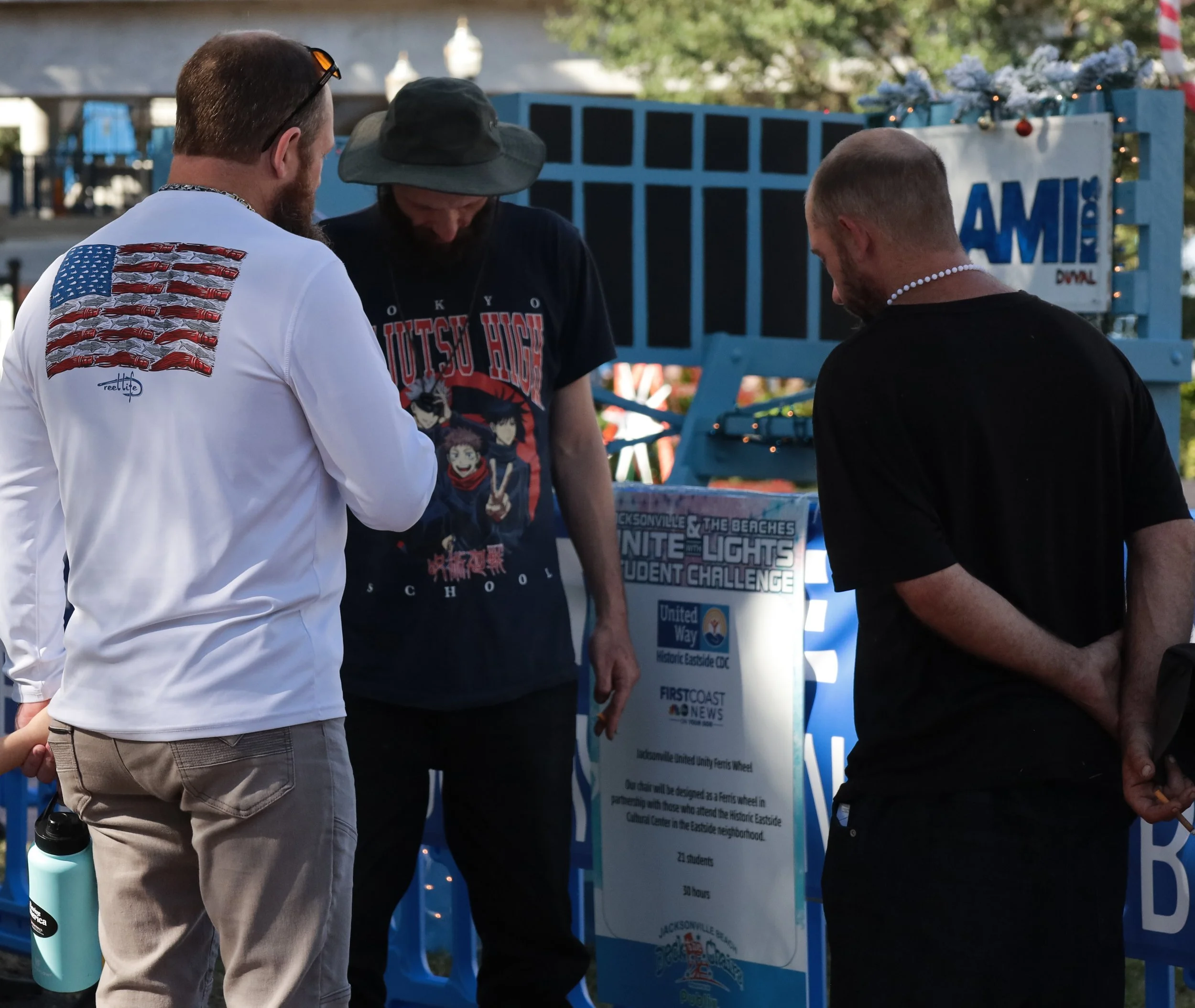 Three men standing and praying near an informational sign at an outdoor event, with a blue wooden structure decorated with Christmas lights in the background.