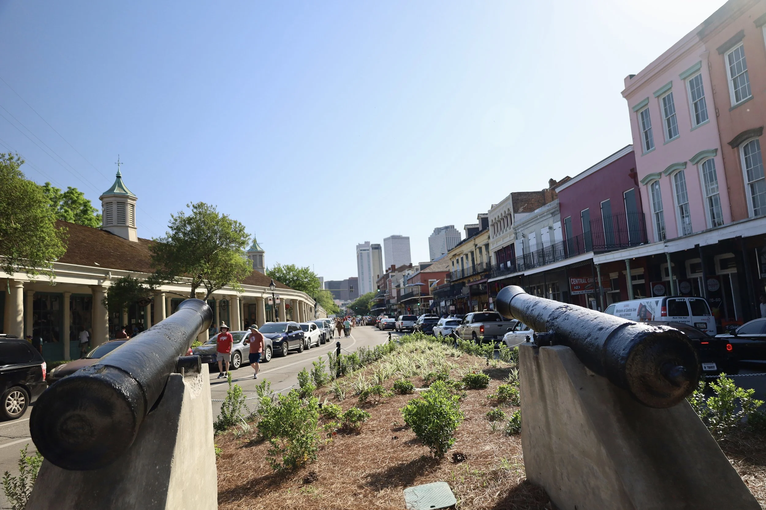 Street view of a historic downtown area with row houses, shops, and cars parked along the street. Cannons in the foreground and modern high-rise buildings in the background.