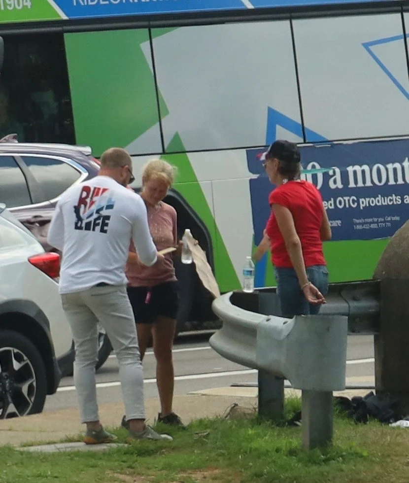 Three people standing near a sidewalk with parked cars and a large bus in the background. The person on the left is wearing a white shirt with red and blue text and beige pants. The middle person is holding a plastic container or bottle, and the pers