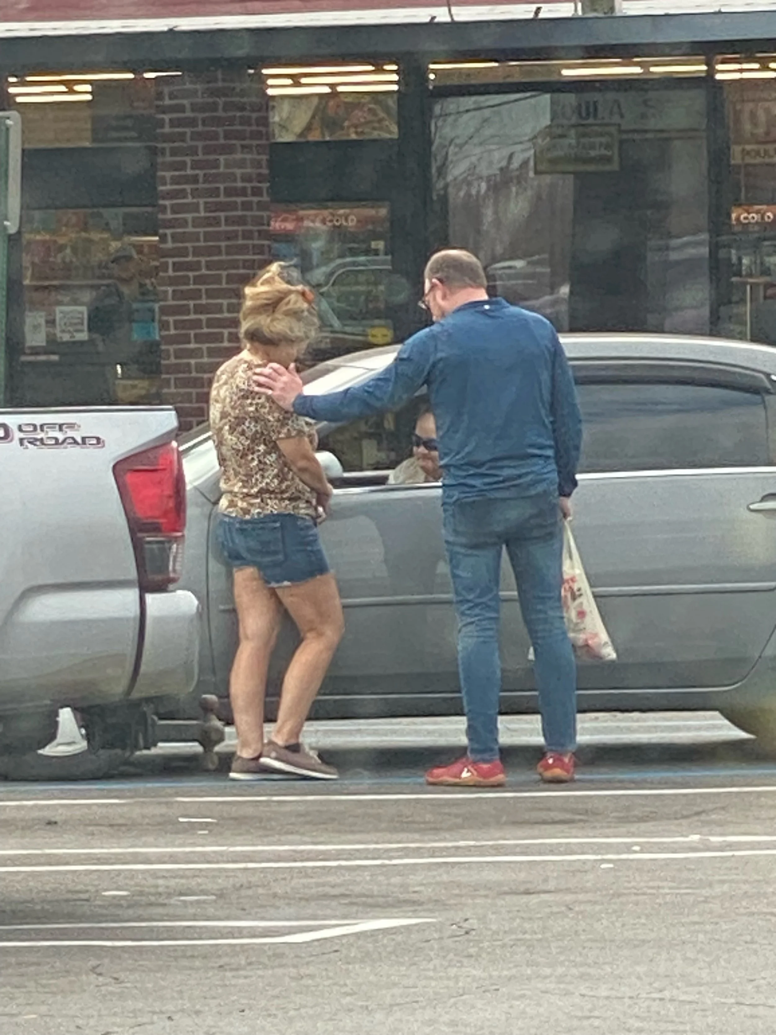 A woman with blond hair wearing a leopard print top and denim shorts standing next to a gray car praying with a man in a blue shirt and jeans outside a store, with another person sitting in the driver's seat of the car.