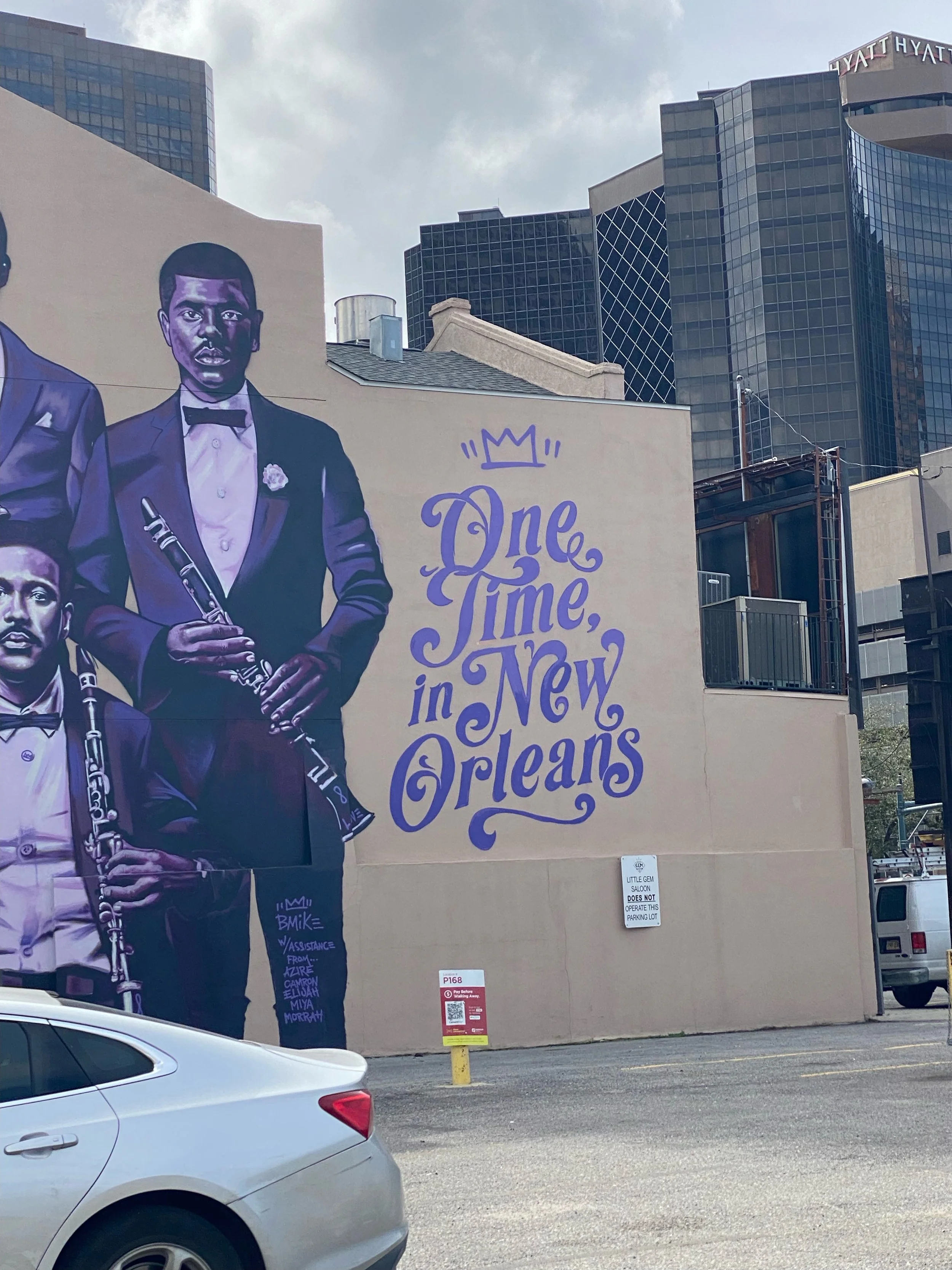 A mural featuring two men in tuxedos holding musical instruments, with the text 'One Time in New Orleans' written in purple script and a small crown above it, on the side of a building in an urban area with tall glass buildings in the background.