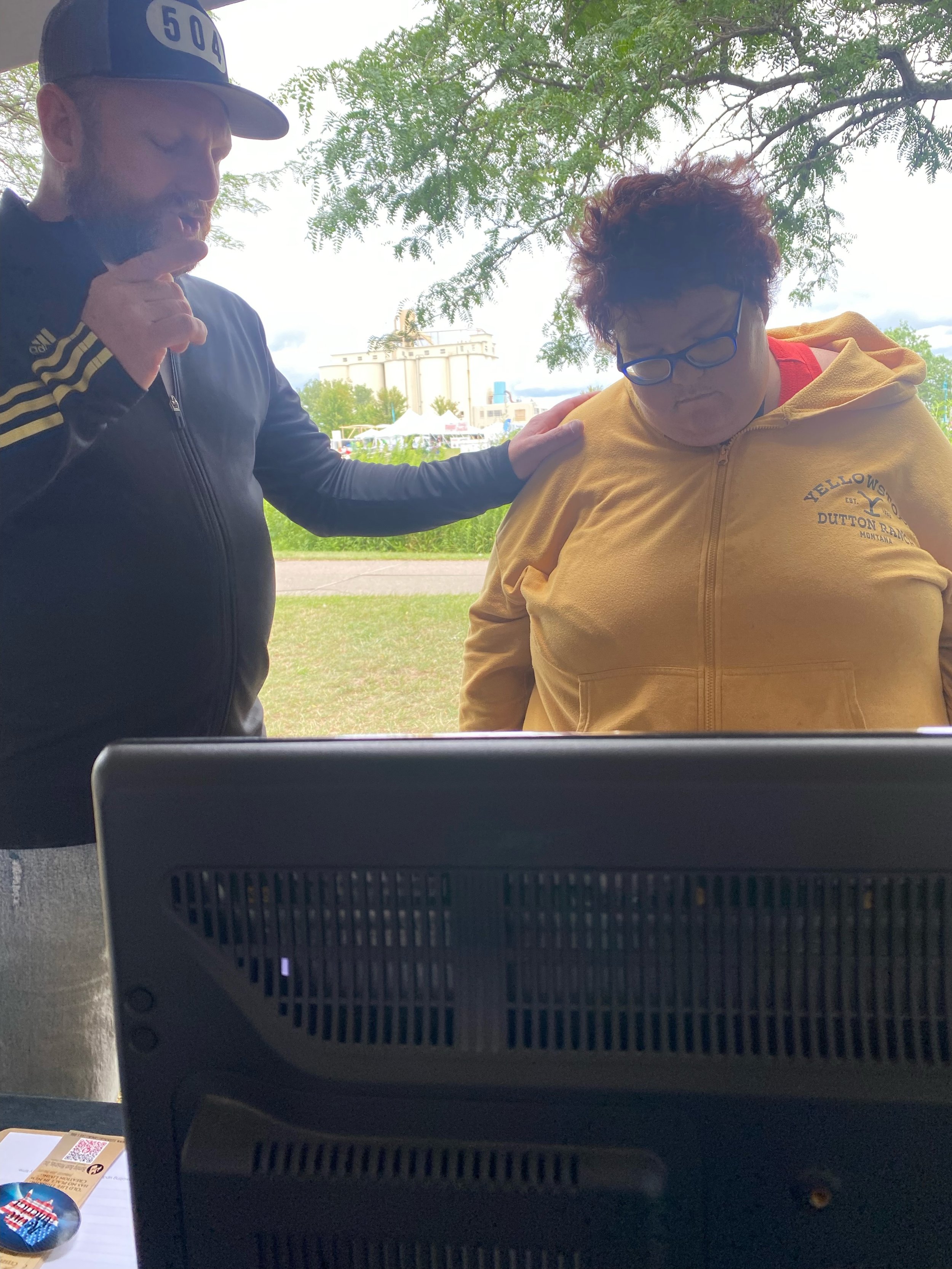 A man and a woman pray outdoors under a tree. The man is wearing a black jacket and baseball cap, and the woman is wearing a yellow hoodie with glasses. The man has his hand on the woman's shoulder praying.