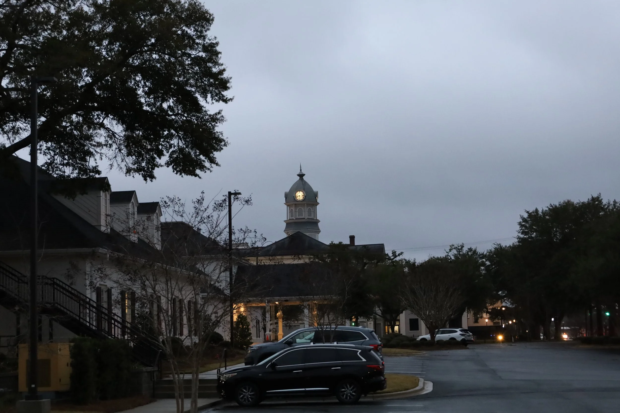 A parking lot with several cars parked. In the background, there are buildings with a clock tower rising above them, trees without leaves, and a cloudy sky.