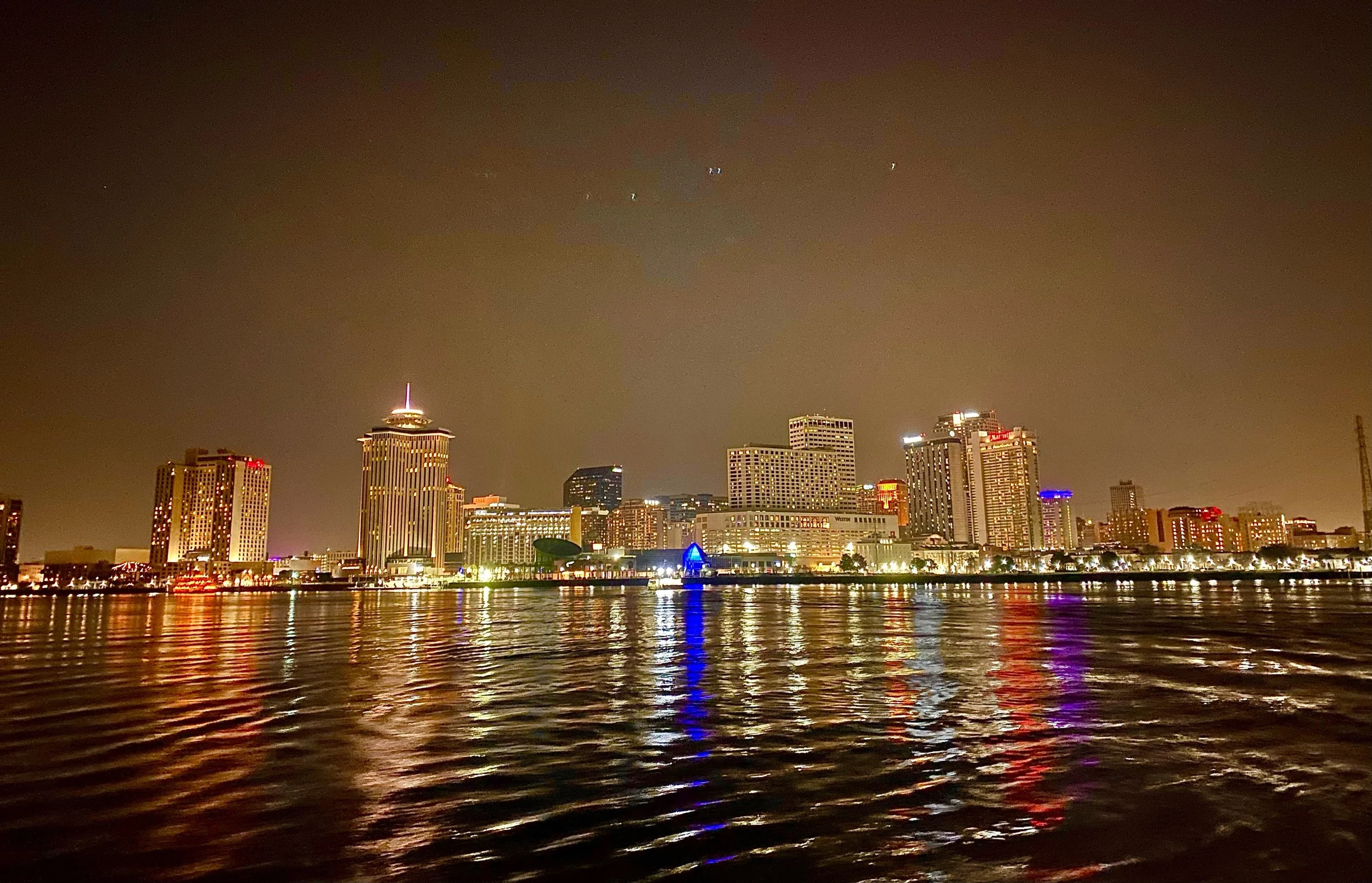 Night view of a city skyline reflecting on water, illuminated skyscrapers, and a cloudy sky.