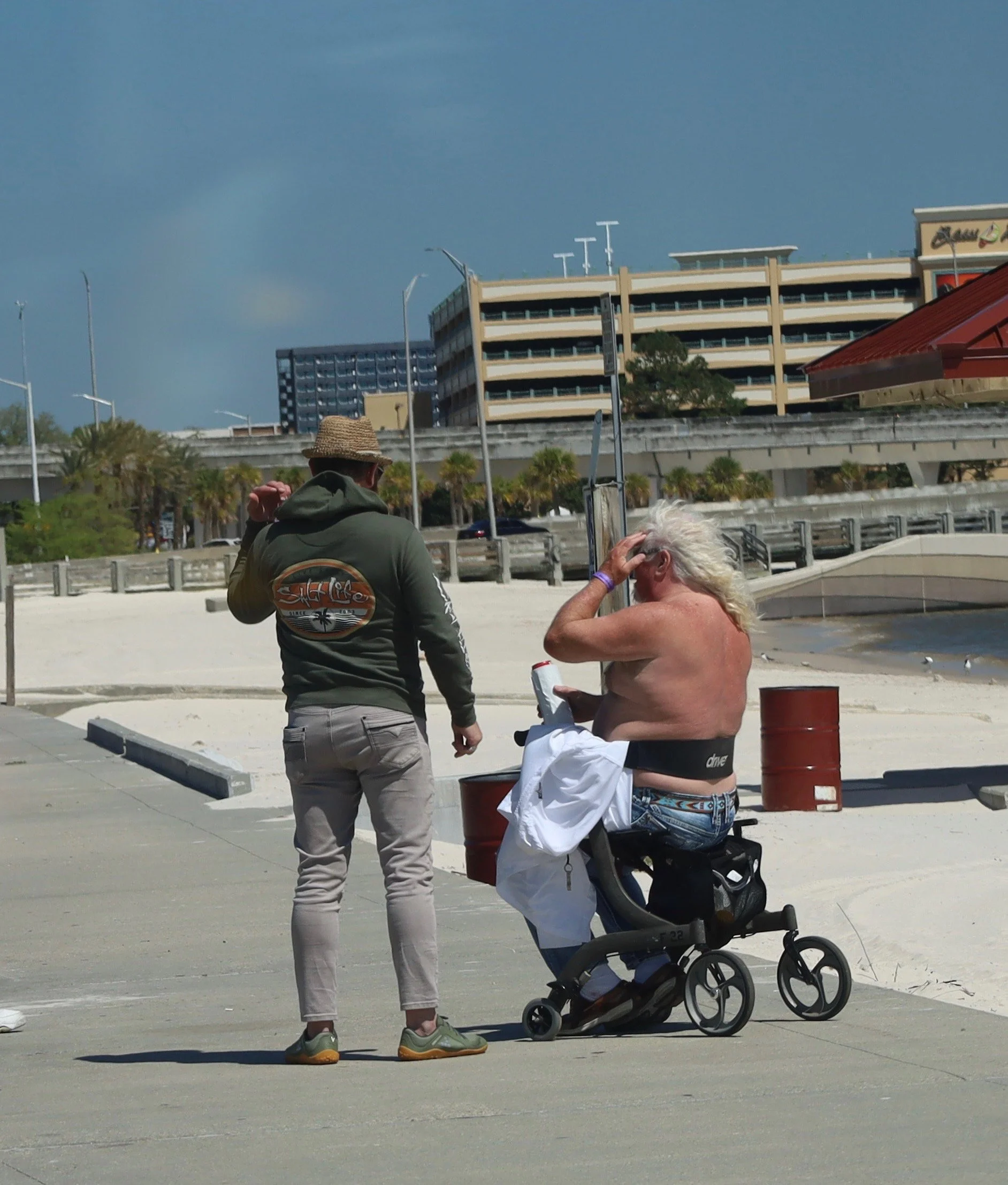 A man with long white hair sitting in a wheelchair on a beach sidewalk, shirtless, talking to a standing man wearing a green hoodie and a straw hat, with a cityscape and sandy beach in the background.