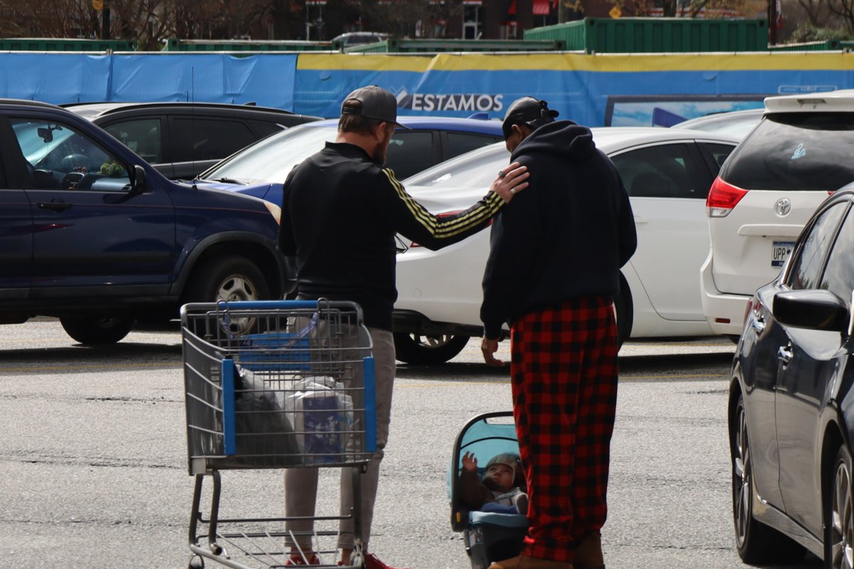 Two men in a parking lot, one with a shopping cart and a baby in a car seat. The man on the left is praying with the other man.