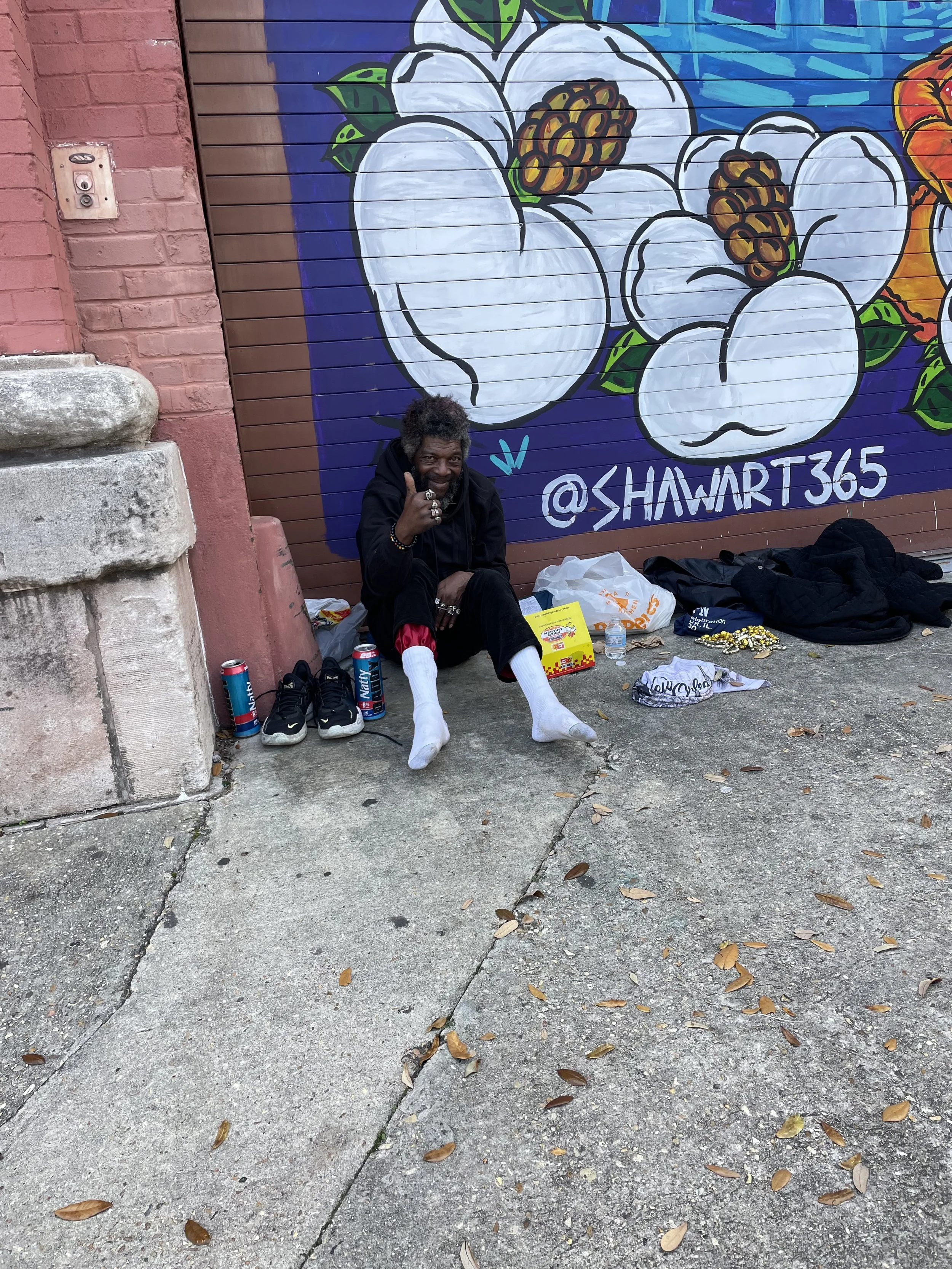 A man sitting on the sidewalk in front of a colorful mural of white flowers with brown centers and green leaves. He is wearing white socks, black pants, and a black hoodie, giving a thumbs-up gesture. There are shoes, cans, a water bottle, and variou