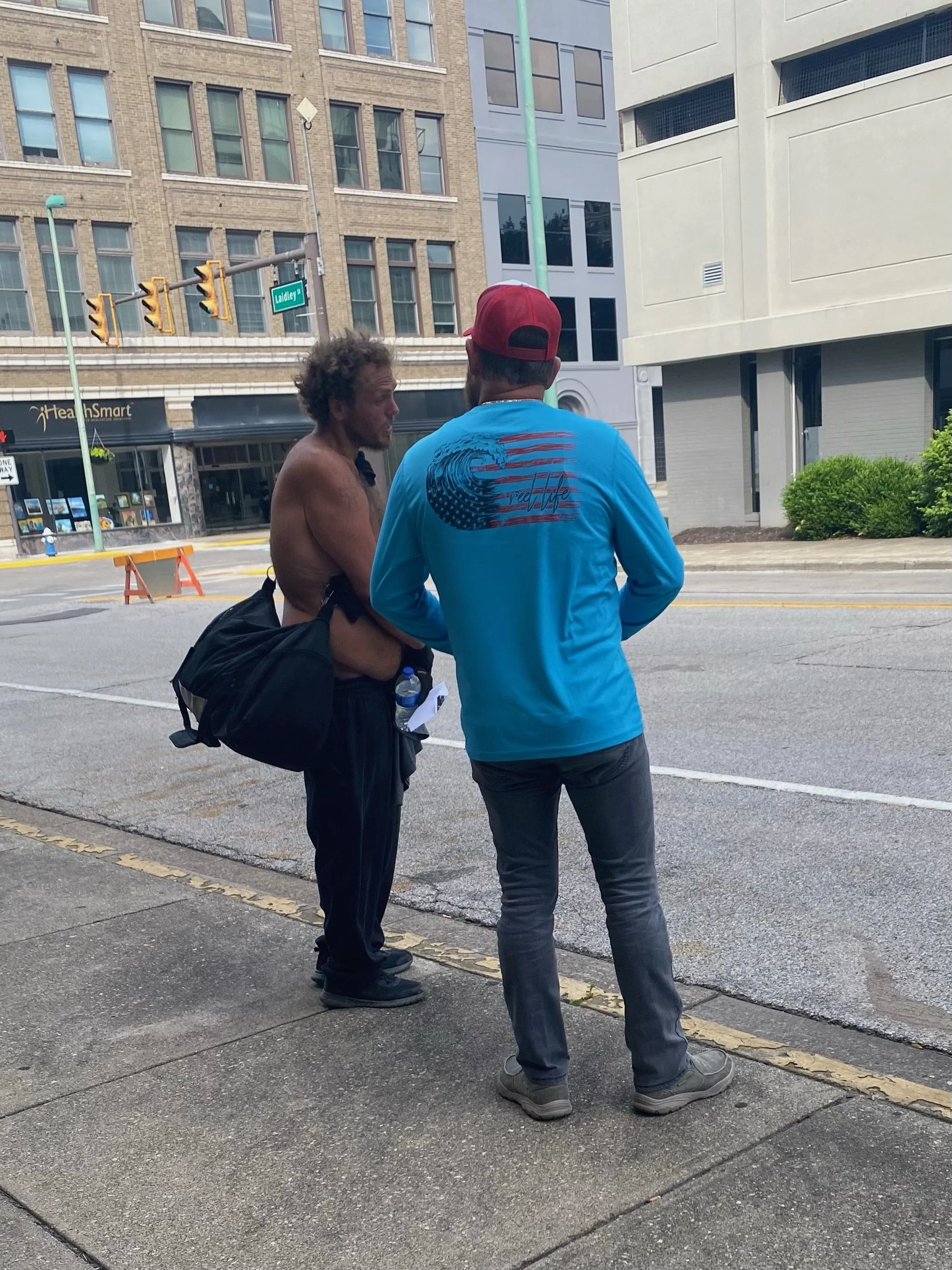 Two men are standing on the sidewalk and talking on a city street. One man is shirtless, facing the other, who is wearing a blue shirt and red cap. There are buildings, traffic lights, and a store sign in the background.