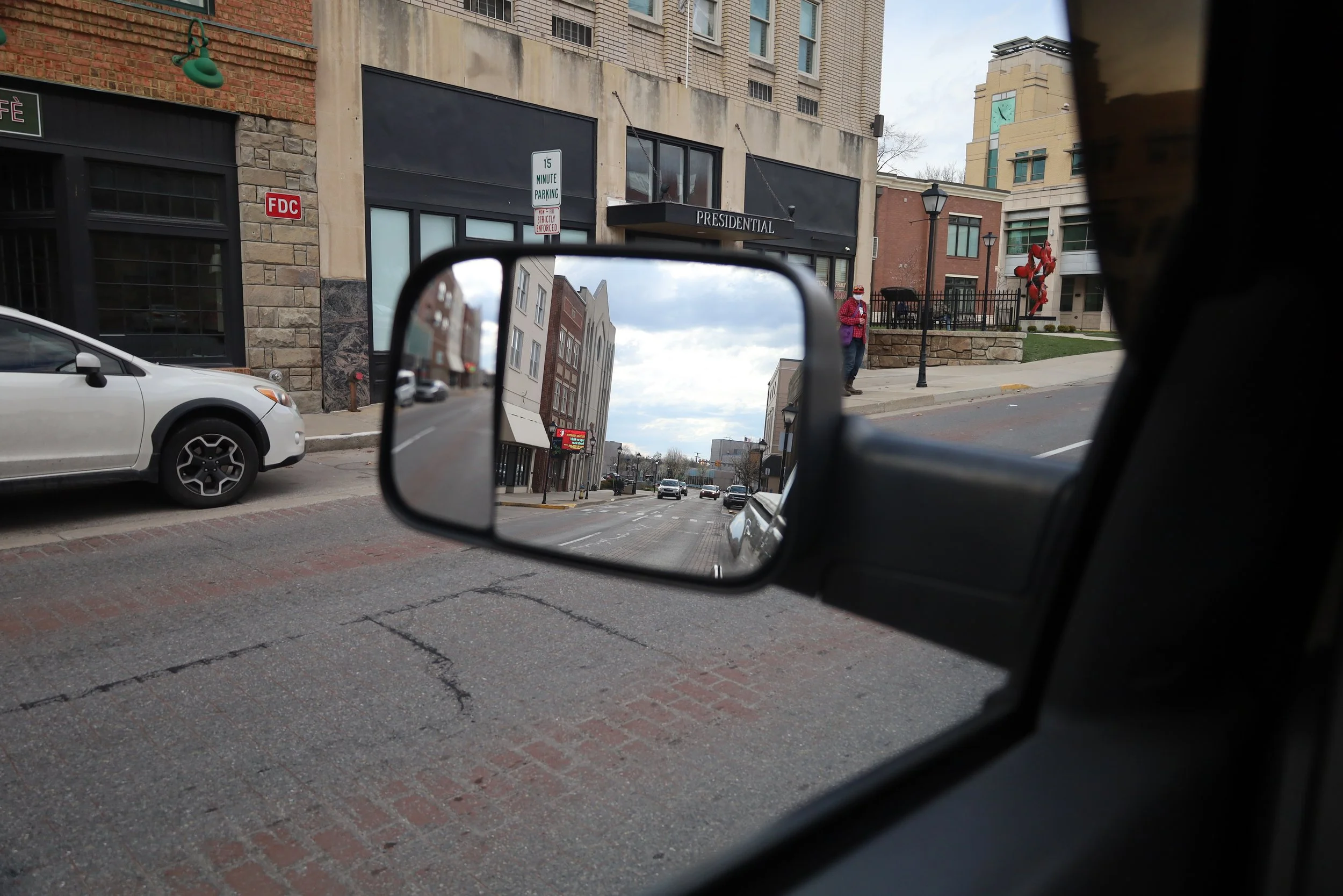 View of a city street seen through a vehicle's side mirror, showing buildings, cars, and pedestrians, including a person in a red outfit and another in a pink jacket praying.