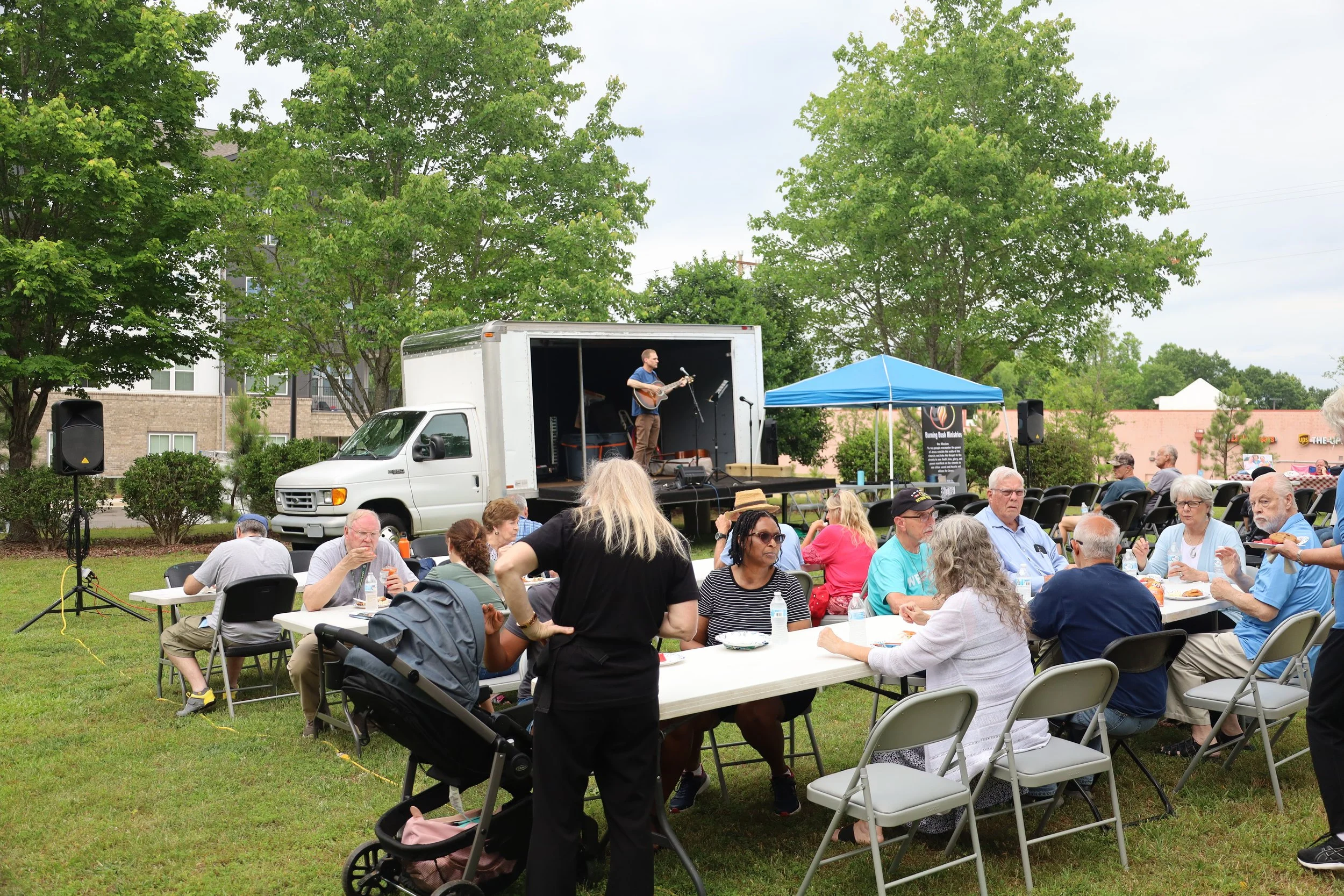 Outdoor gathering with people seated at tables enjoying food, watching a worship session on a small stage with a guitarist, under a blue canopy, in a park-like setting with trees and buildings in the background.